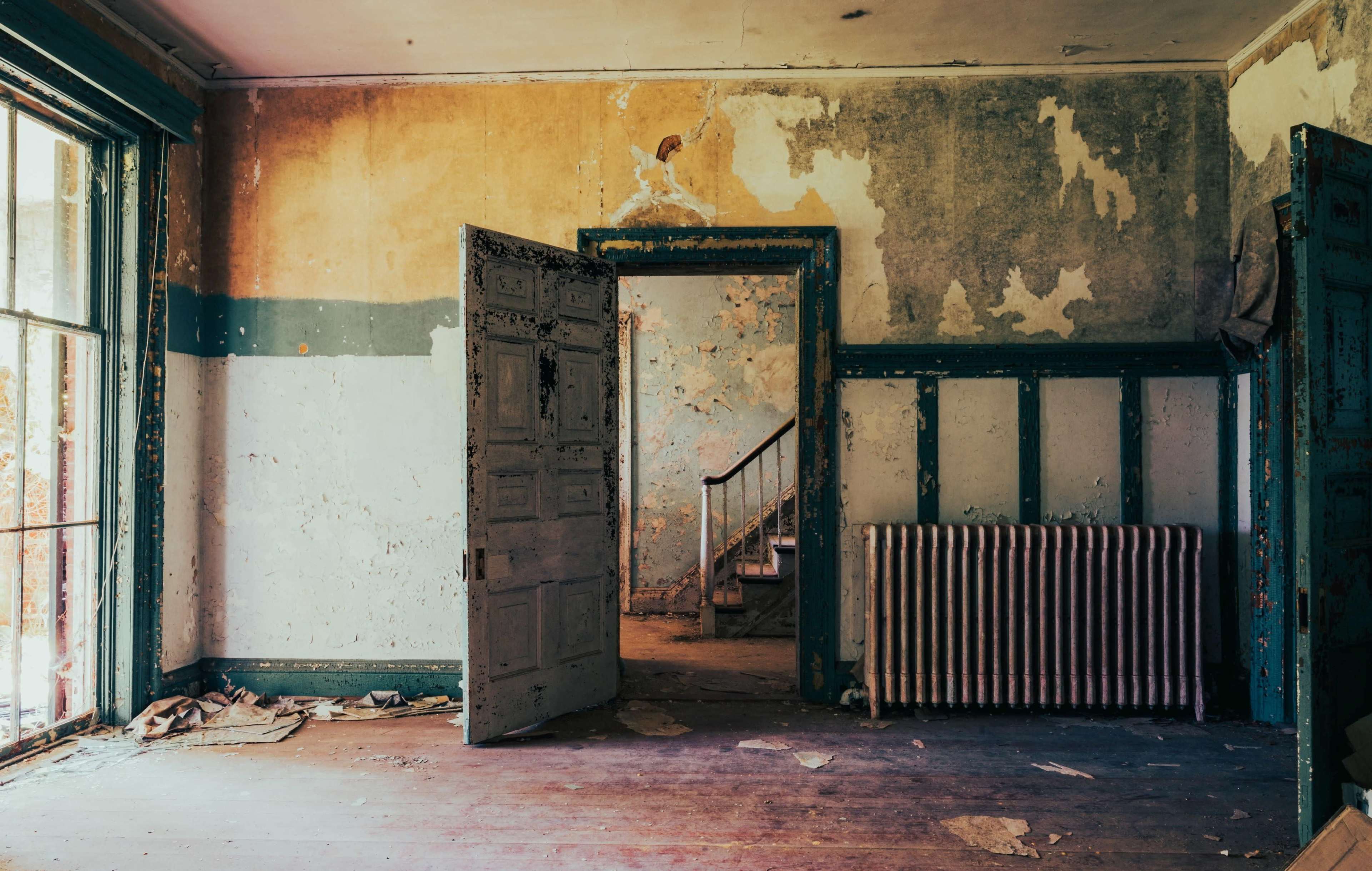 An empty room with peeling paint, an open door leading to a staircase, and a radiator against the wall.