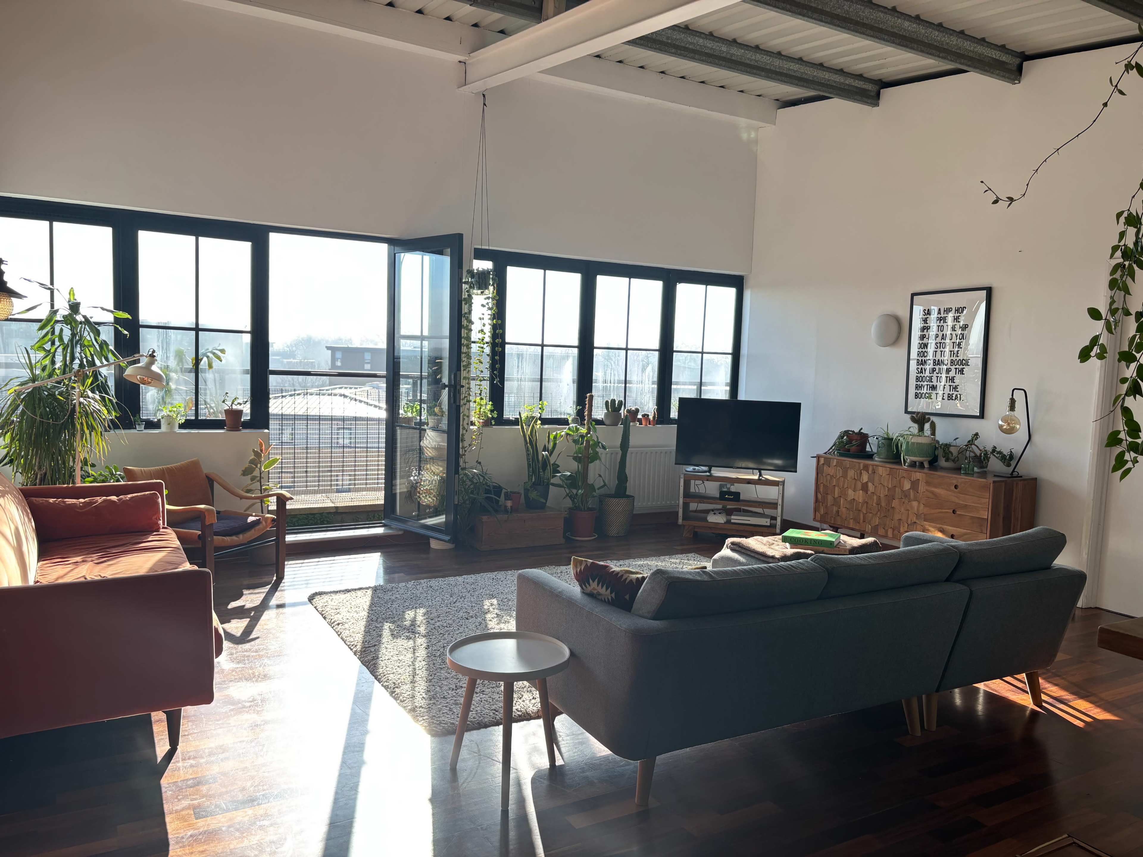 A spacious living room features large windows, a light rug, a gray sofa, wooden furniture, and various indoor plants.