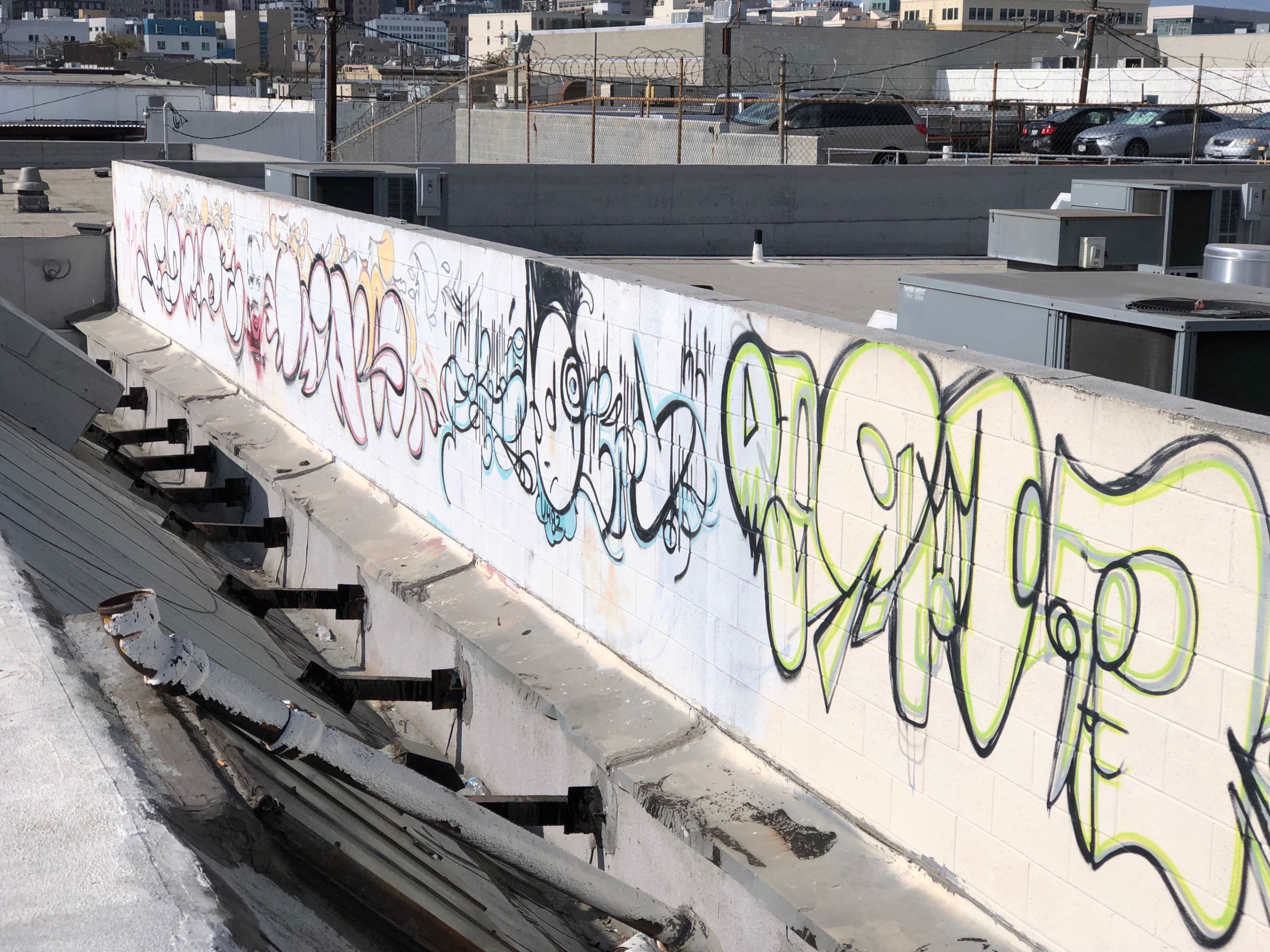 A long wall covered in graffiti, with various colorful tags and letters displayed against a backdrop of rooftops and distant buildings.