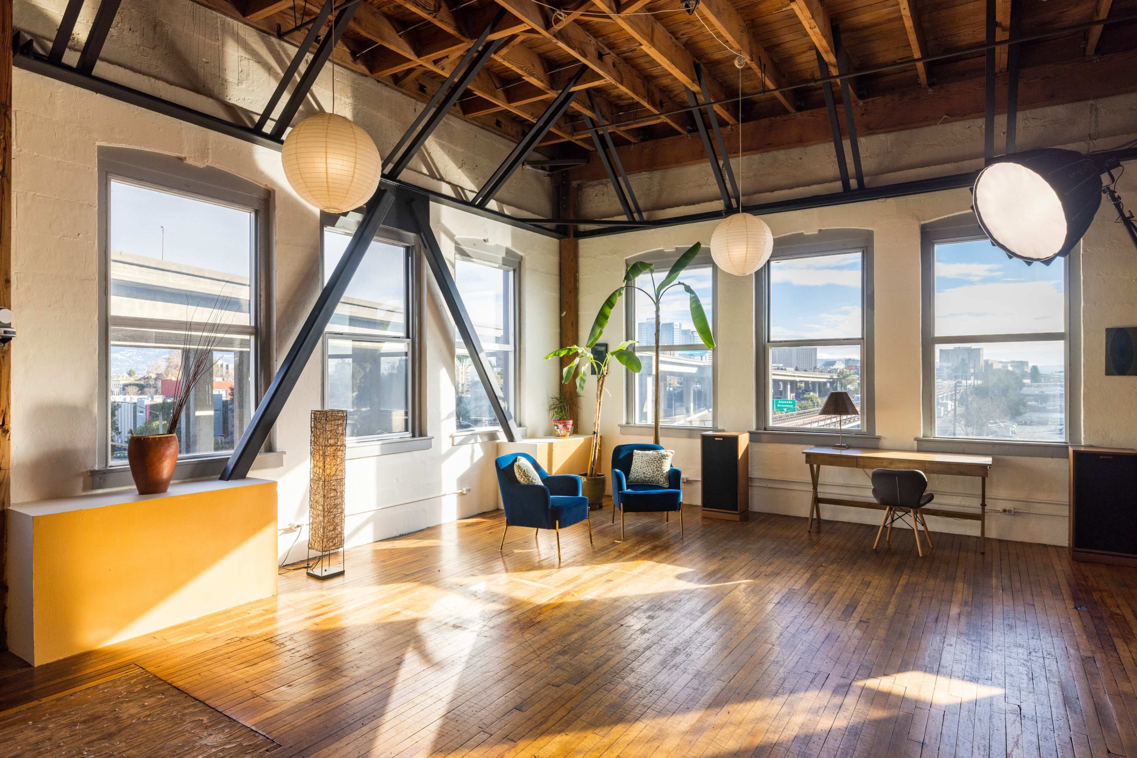 A bright, spacious room features large windows, wooden floors, and modern furniture, including two chairs, a desk, and decorative elements.
