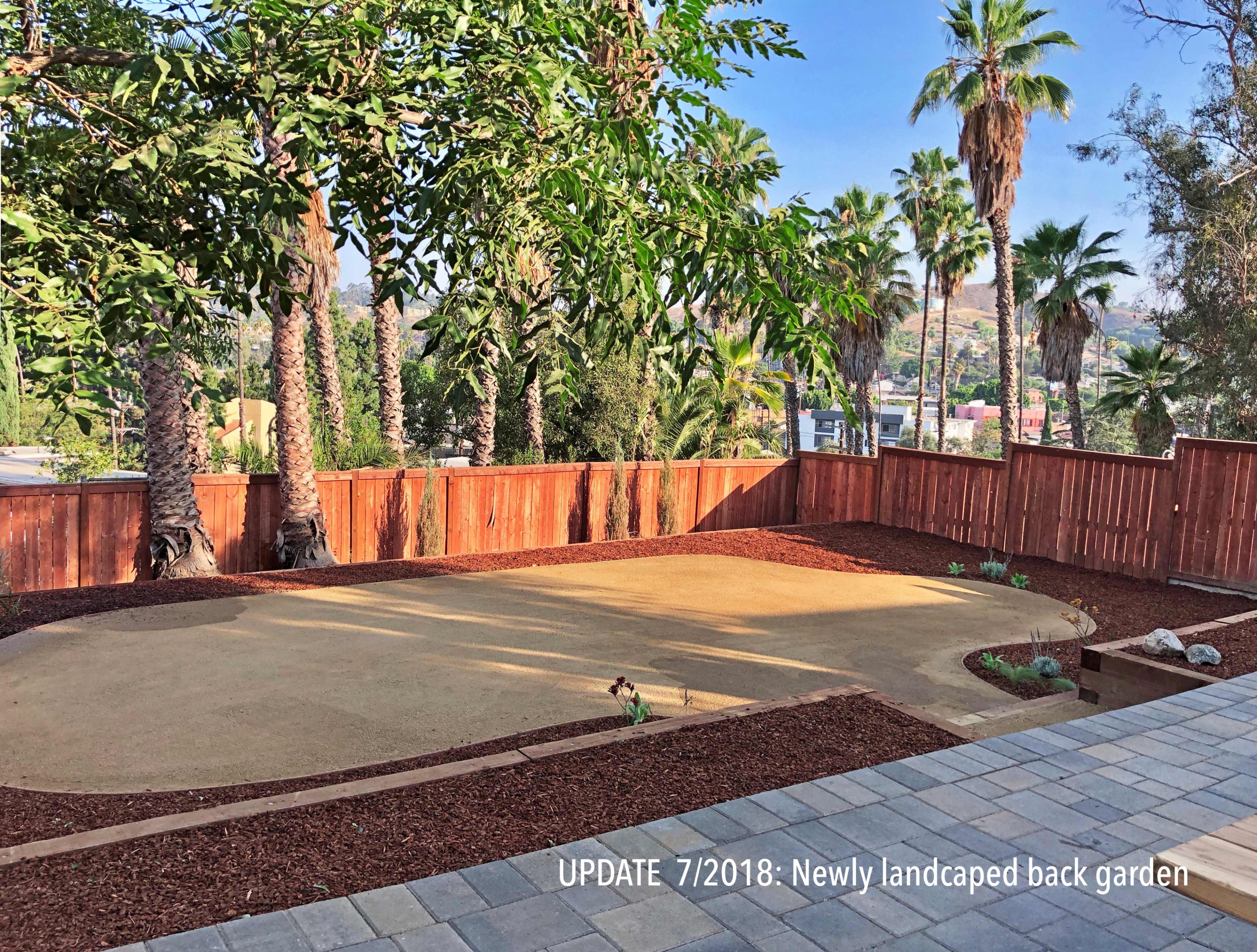 A newly landscaped back garden featuring a sandy area surrounded by mulch, palm trees, and a wooden fence.