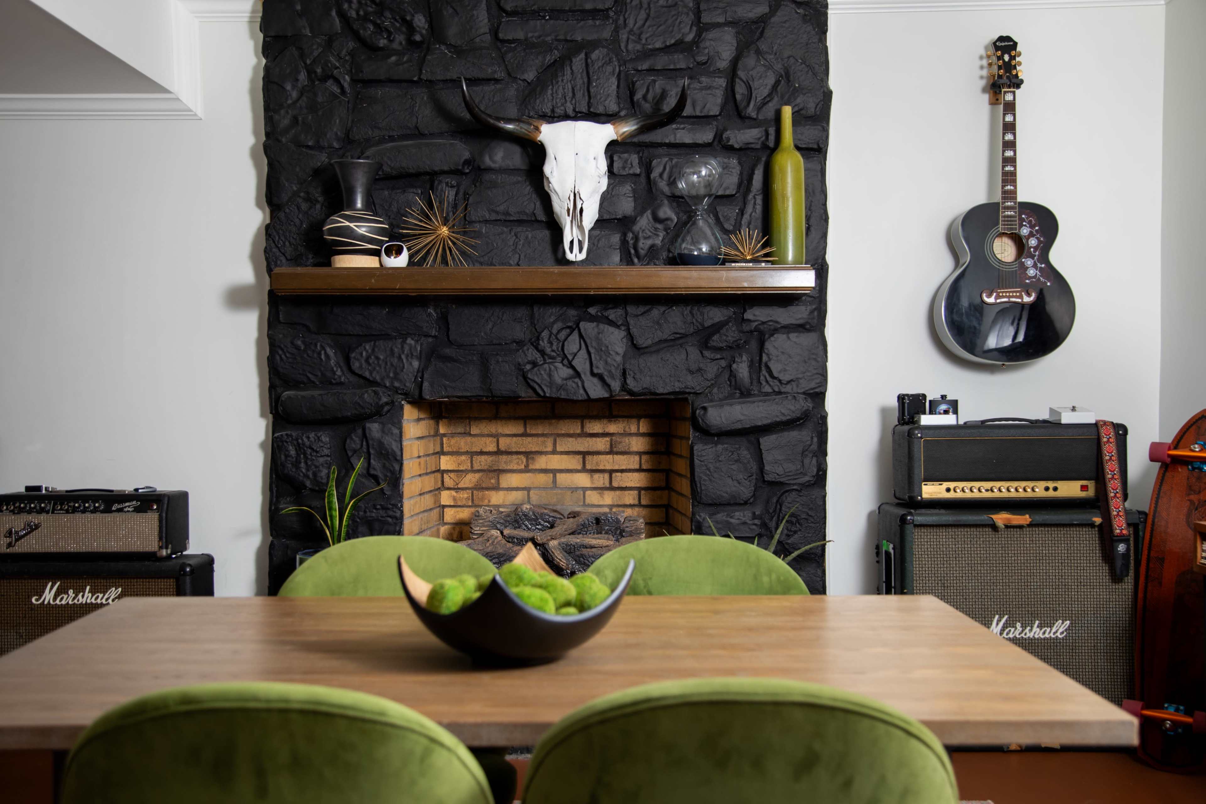 A dining area features a wooden table with green chairs in front of a black stone fireplace adorned with a skull, shelf decor, and musical instruments on the walls.