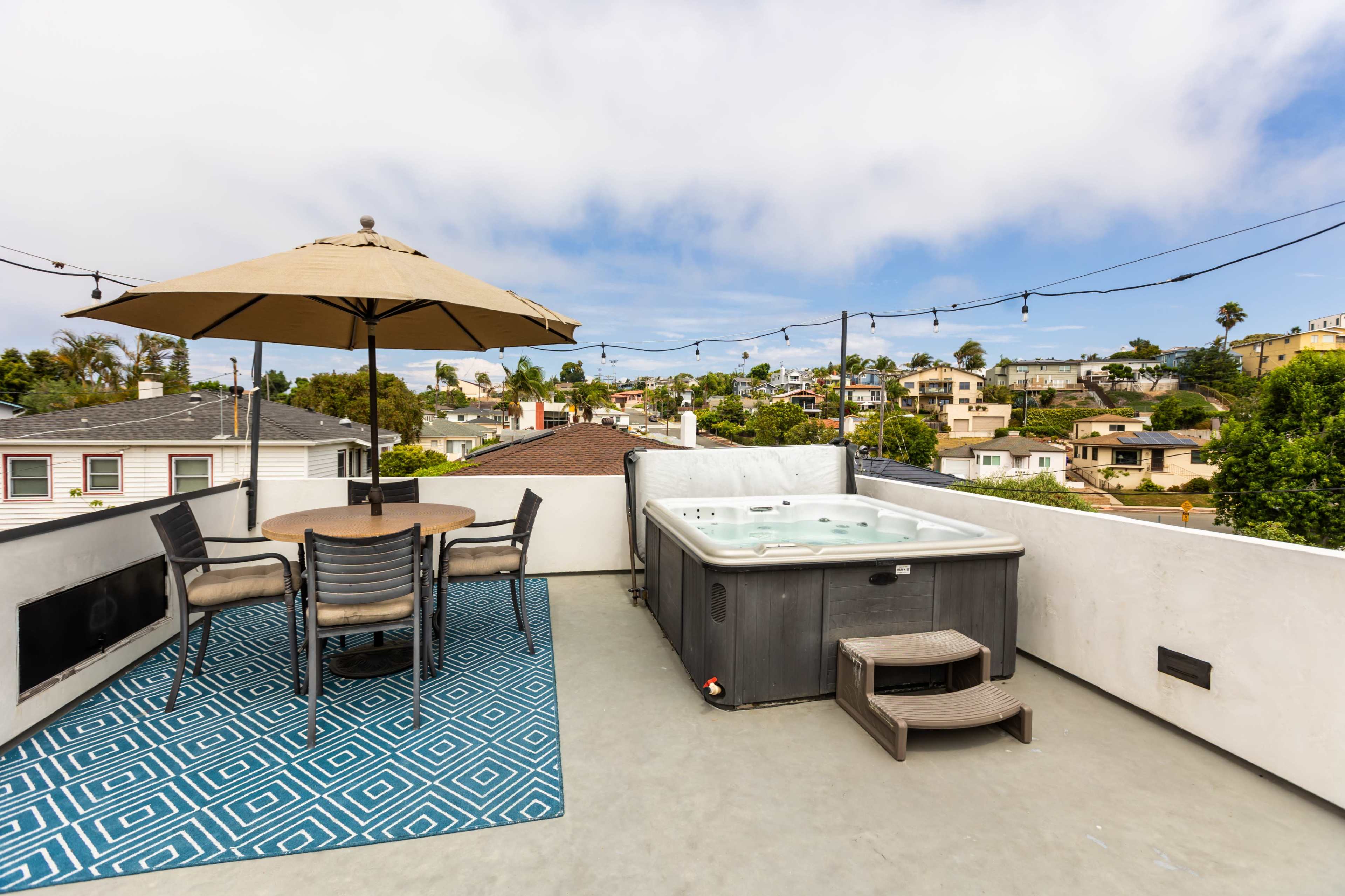 The rooftop features a hot tub, a dining table with chairs, and an umbrella, overlooking a neighborhood with houses and trees.