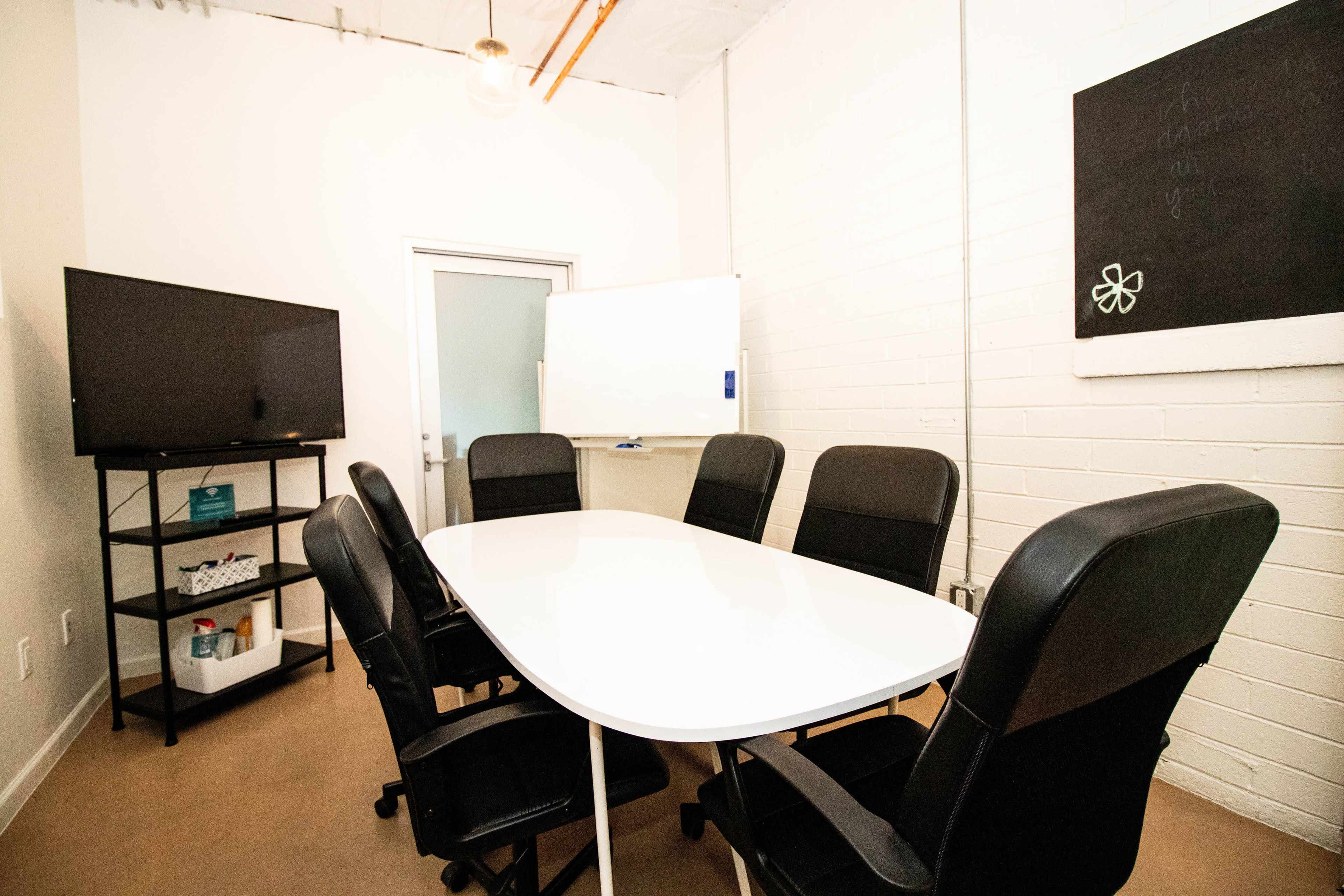 A conference room features a white oval table surrounded by six black ergonomic chairs, with a television and a whiteboard on the walls.