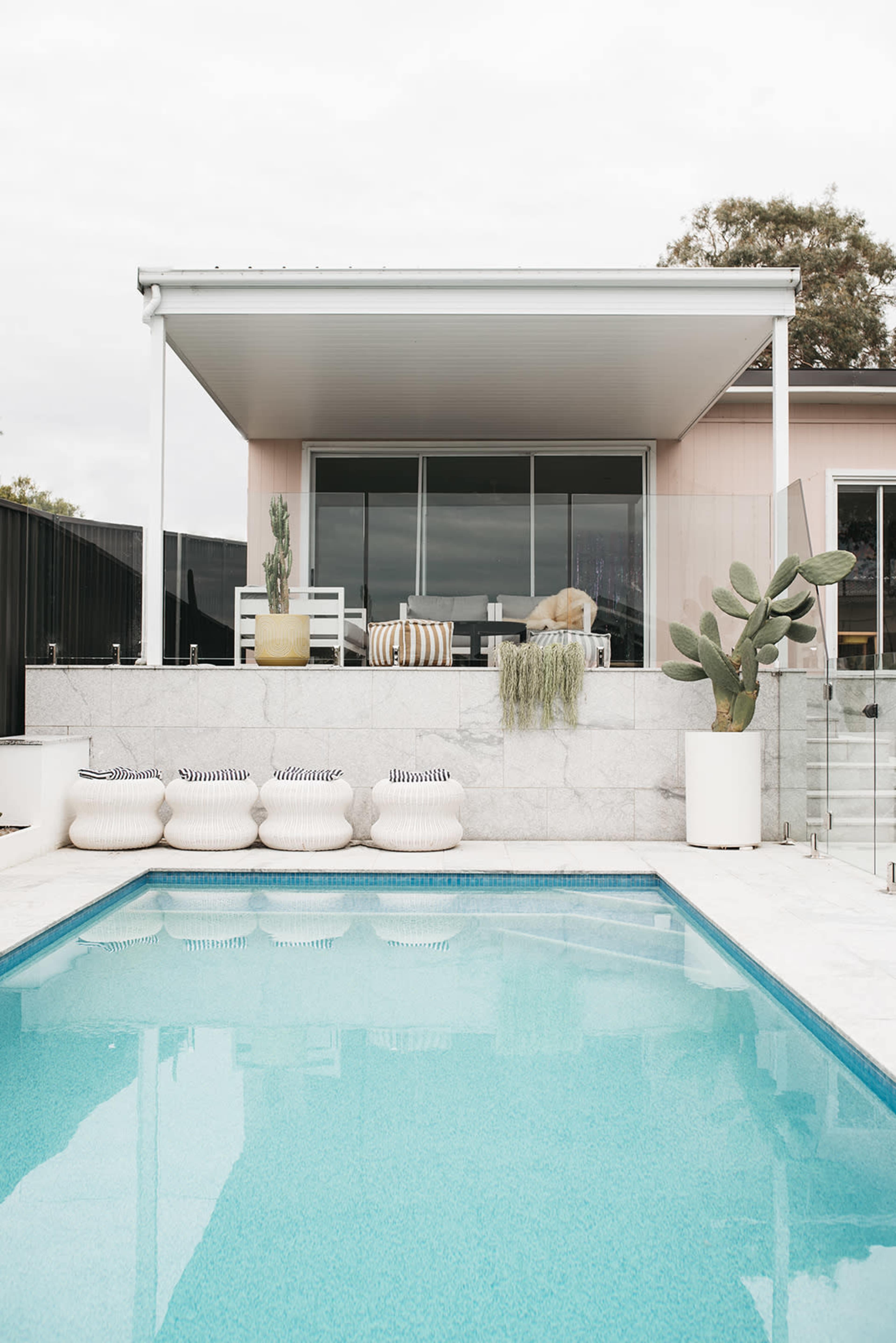 A modern outdoor space features a swimming pool in the foreground, with a seating area and potted plants visible behind a glass wall.