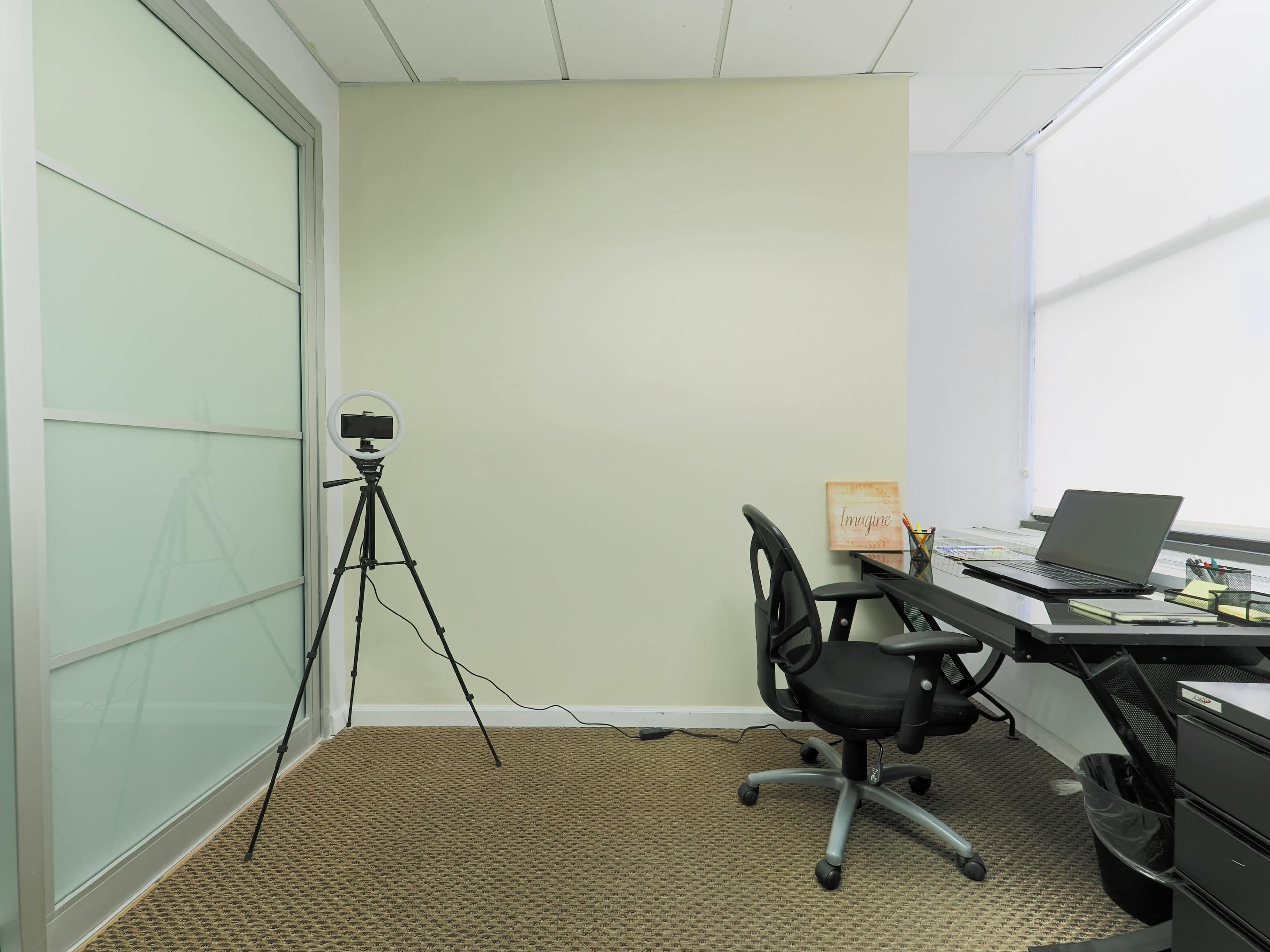 A small office space featuring a desk with a laptop, a swivel chair, a tripod holding a camera, and a wall with large windows.