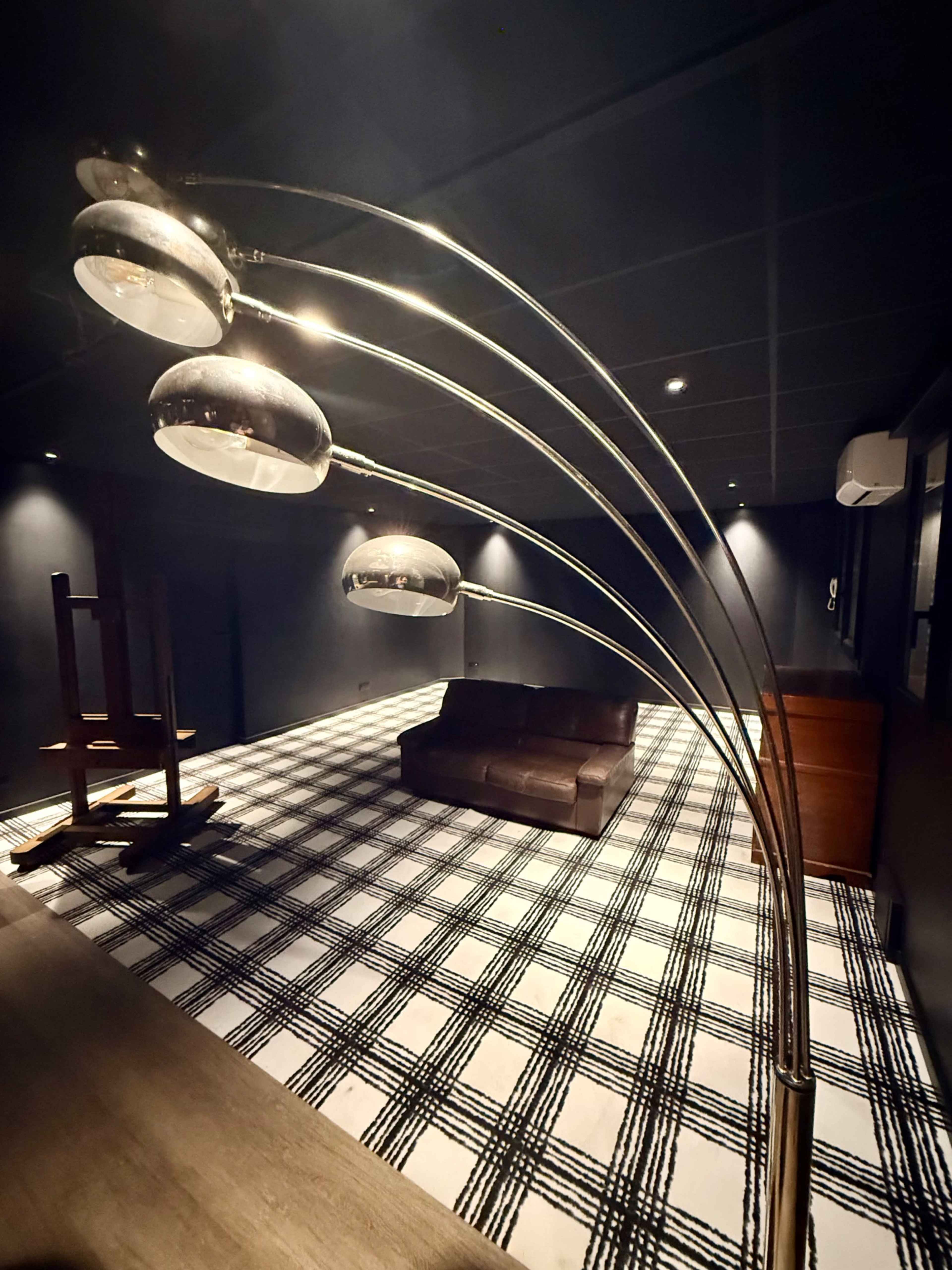 A modern floor lamp with multiple shades stands in a dimly lit room featuring a checkered floor and a brown sofa.