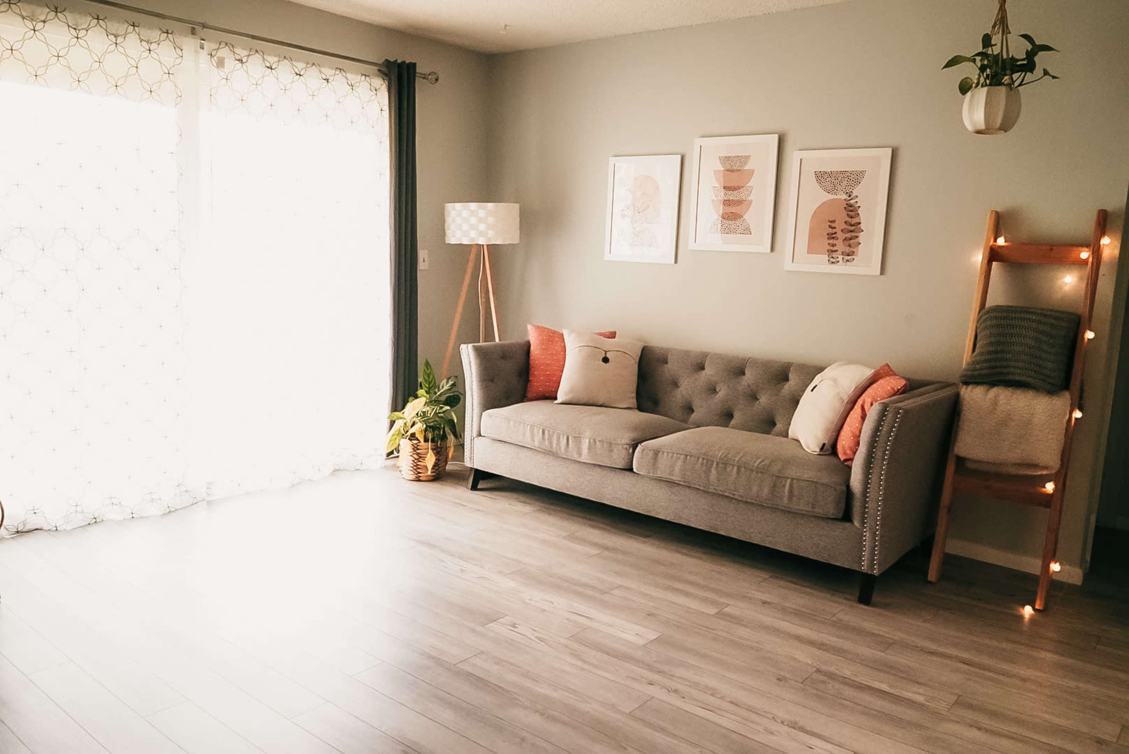A gray tufted sofa with decorative cushions is positioned in a well-lit room featuring framed artwork, a floor lamp, and a wooden ladder displaying a blanket.