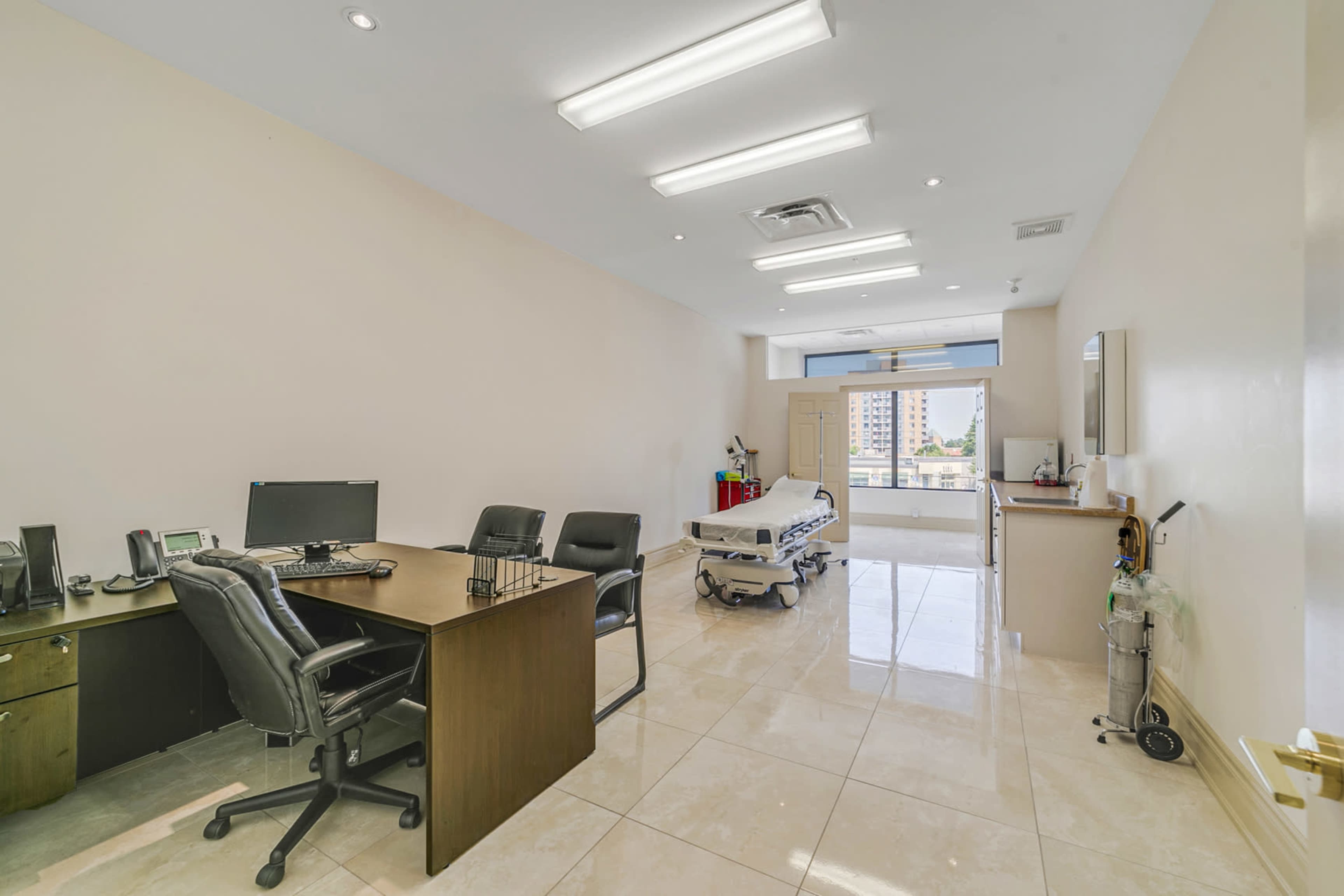 The image shows a clean and well-lit medical examination room featuring a desk with office chairs, a medical examination bed, and a window overlooking an exterior view.