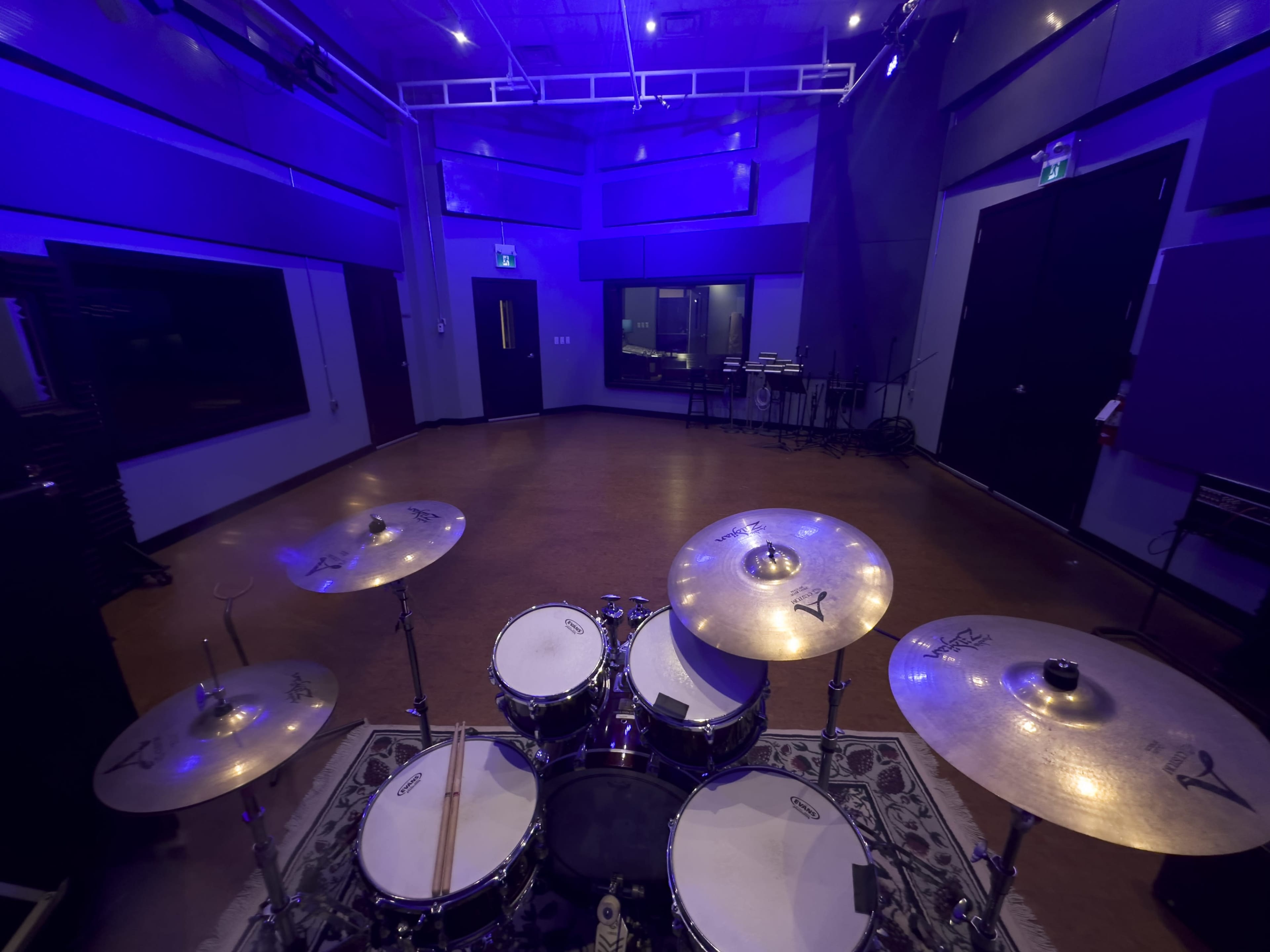 The image shows a view of a music studio from behind a drum set, featuring purple lighting and soundproof walls.