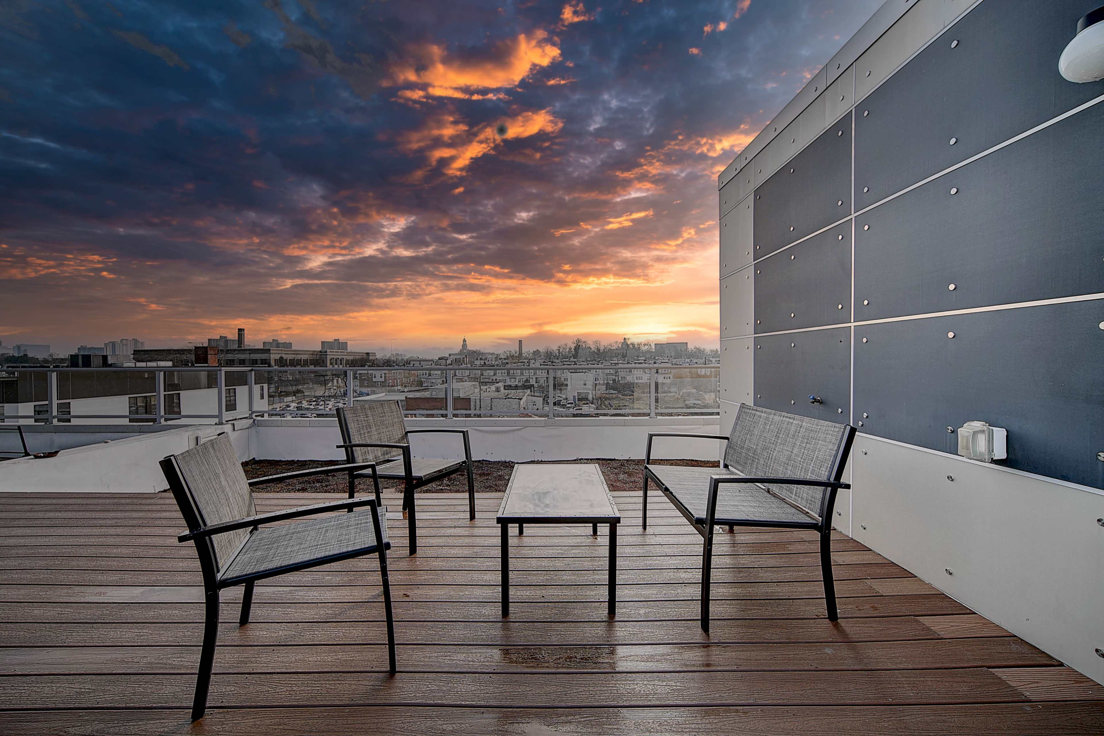 A rooftop terrace features four chairs and a coffee table, with a sunset skyline visible in the background.