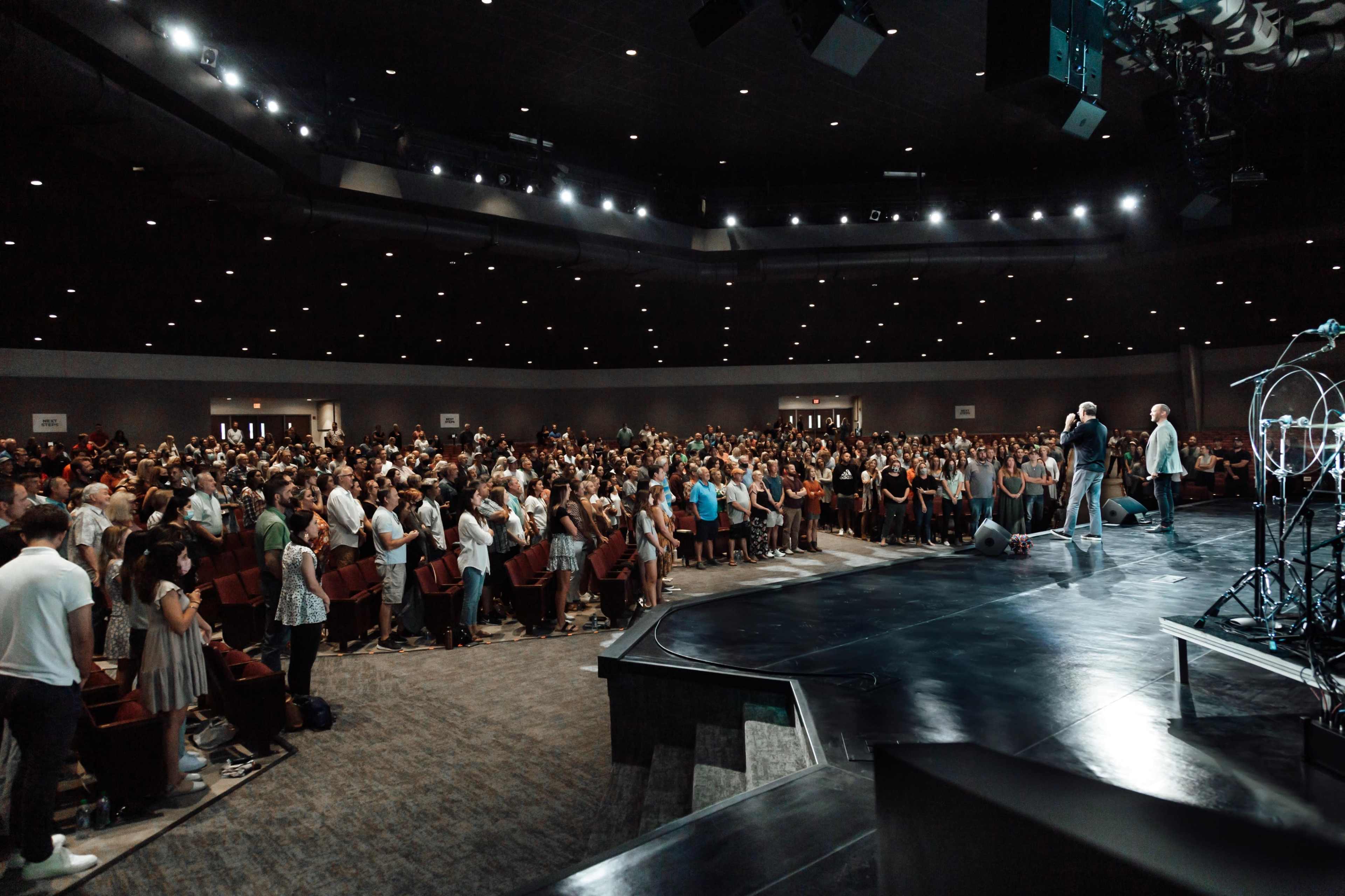 A large audience stands in a dimly lit auditorium, facing a stage where speakers are addressing them.