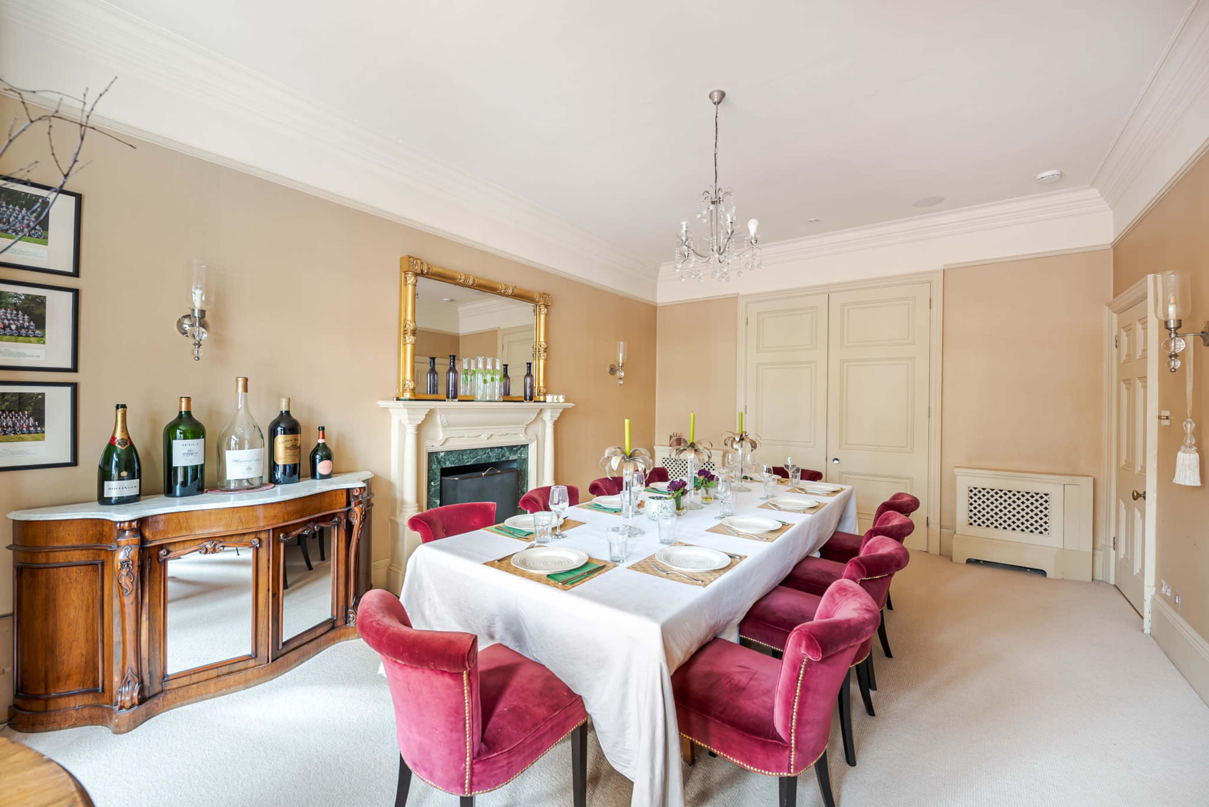 A long dining table is set with plates and glassware, surrounded by red chairs, in a well-lit room with a mirror and a decorative fireplace.