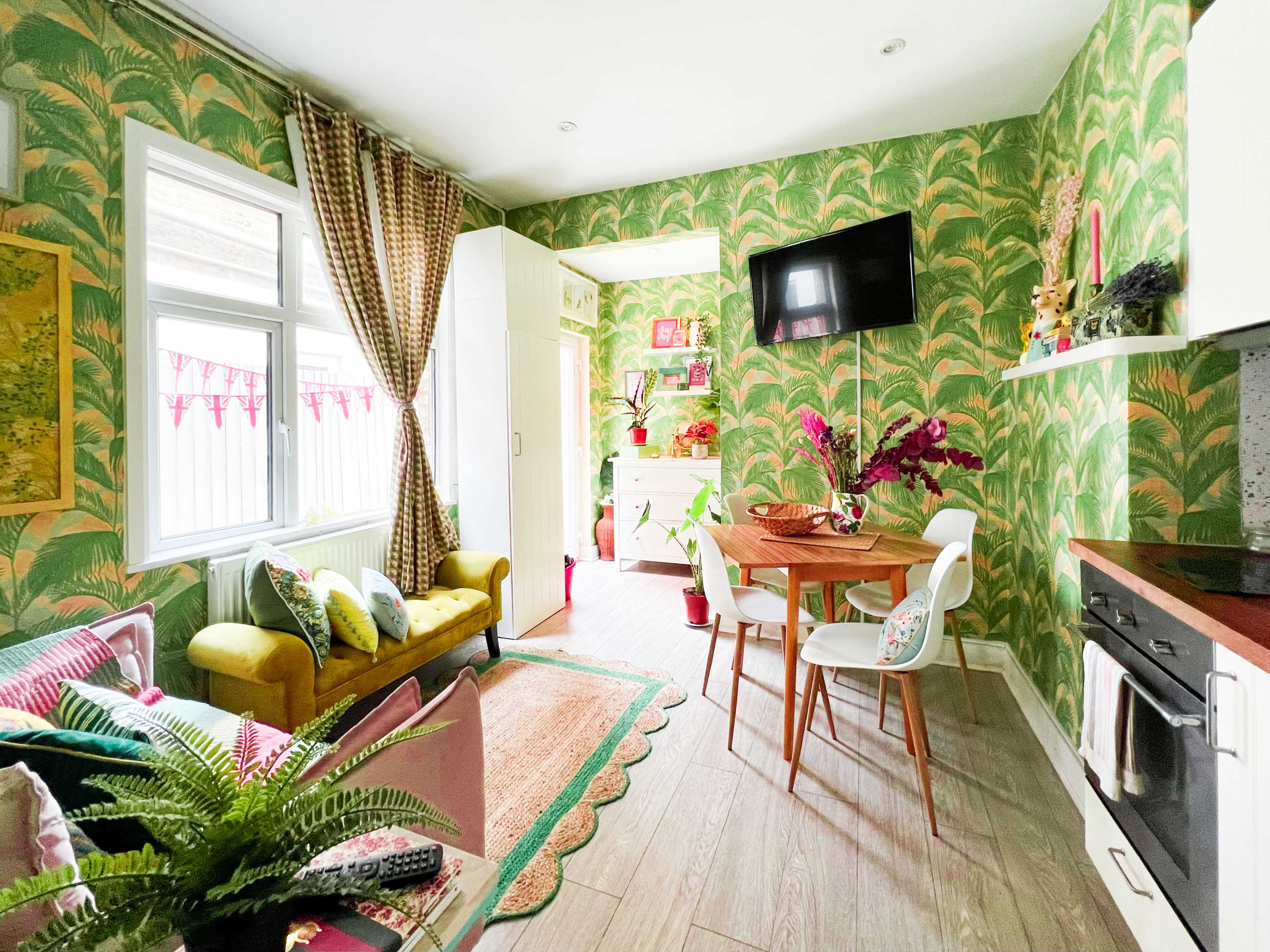The image shows a small, brightly decorated kitchen and dining area with green floral wallpaper, a round dining table, and a cozy seating arrangement.