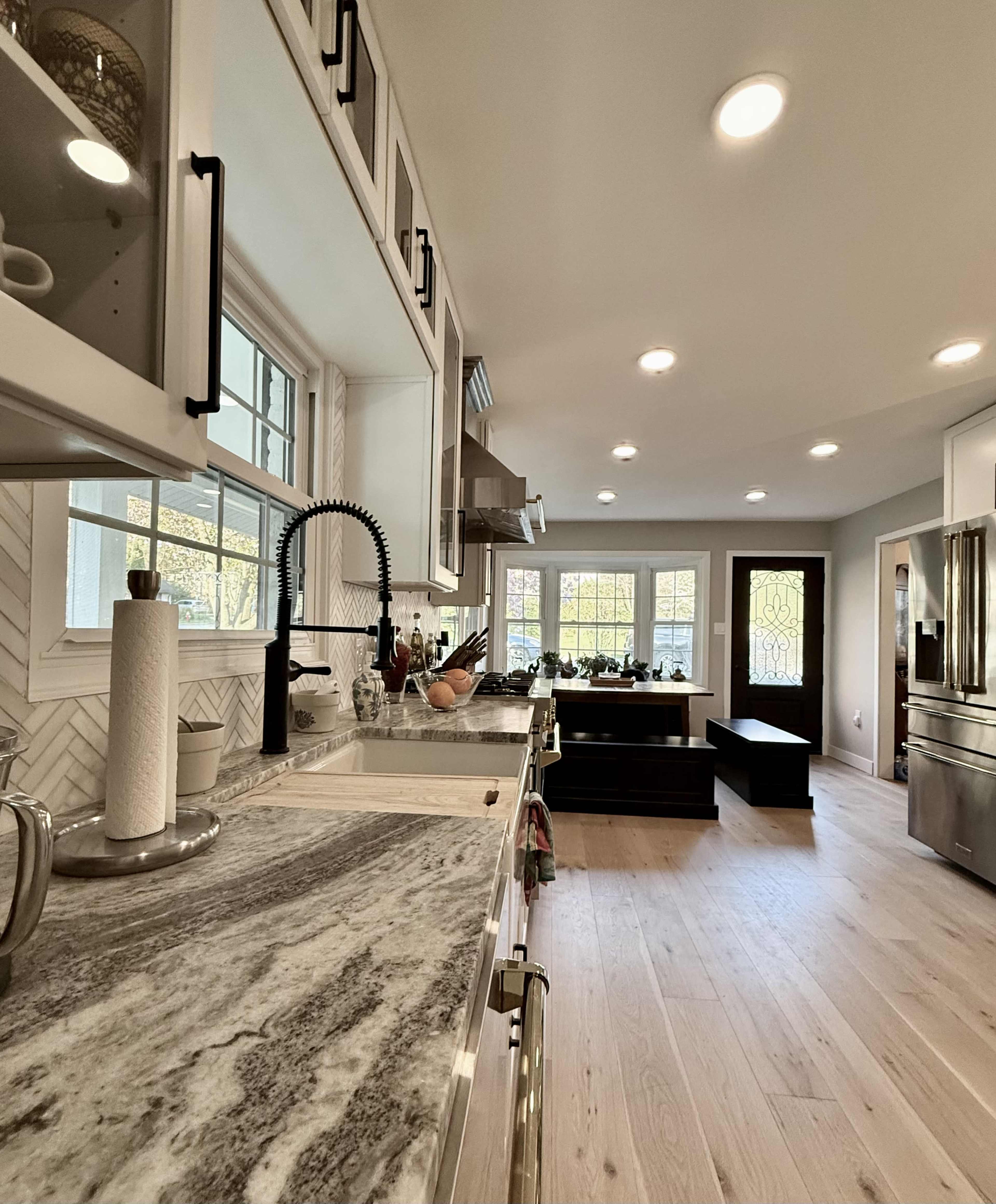The image shows a modern kitchen with a marble countertop, stainless steel appliances, and a view of large windows that let in natural light.