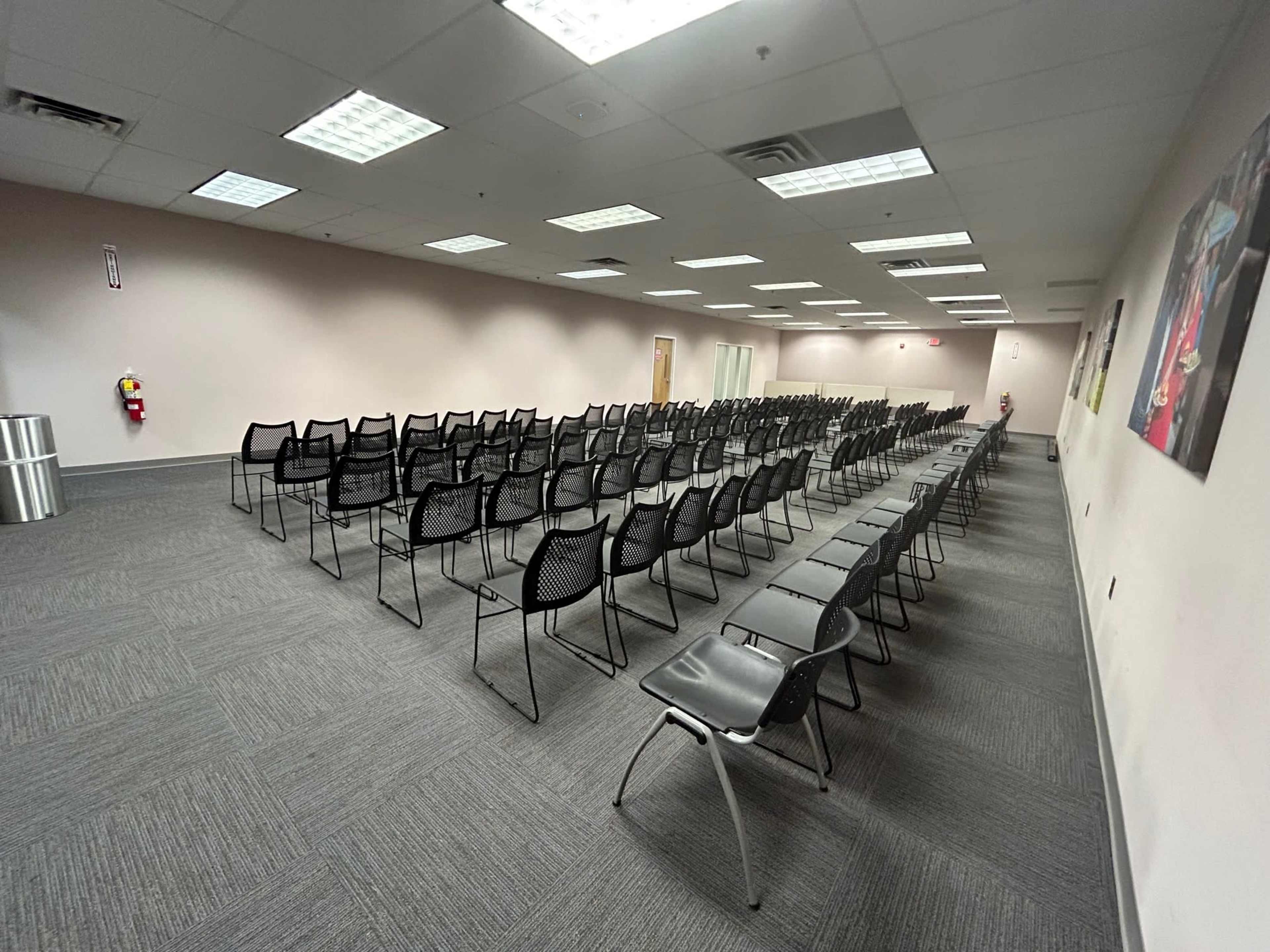 The image shows a large meeting room arranged with rows of black chairs facing the front, with beige walls and fluorescent lighting.
