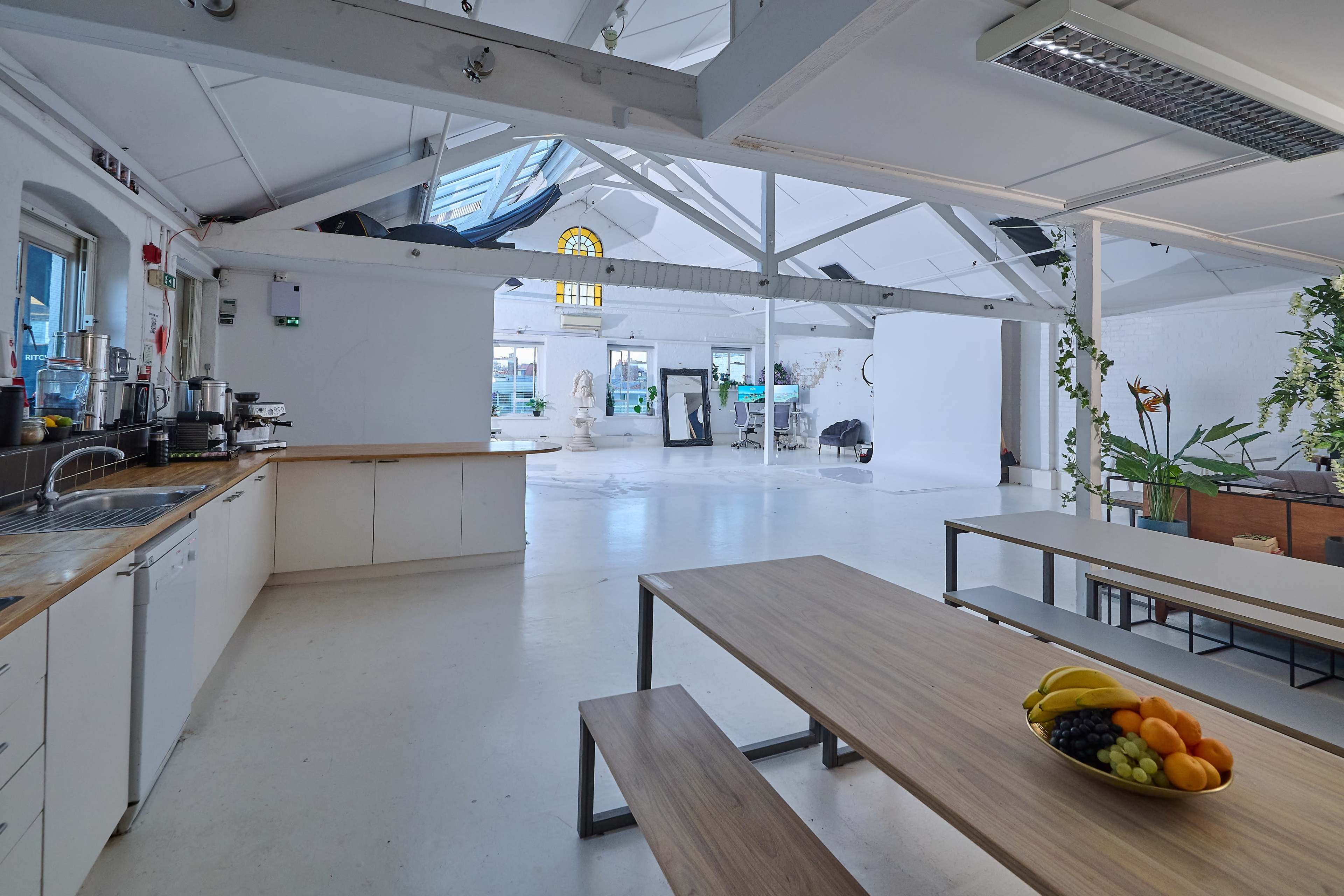 The image shows a spacious, well-lit loft-style kitchen with a wooden dining table and a bowl of fruit, adjacent to a large open area used for photography.