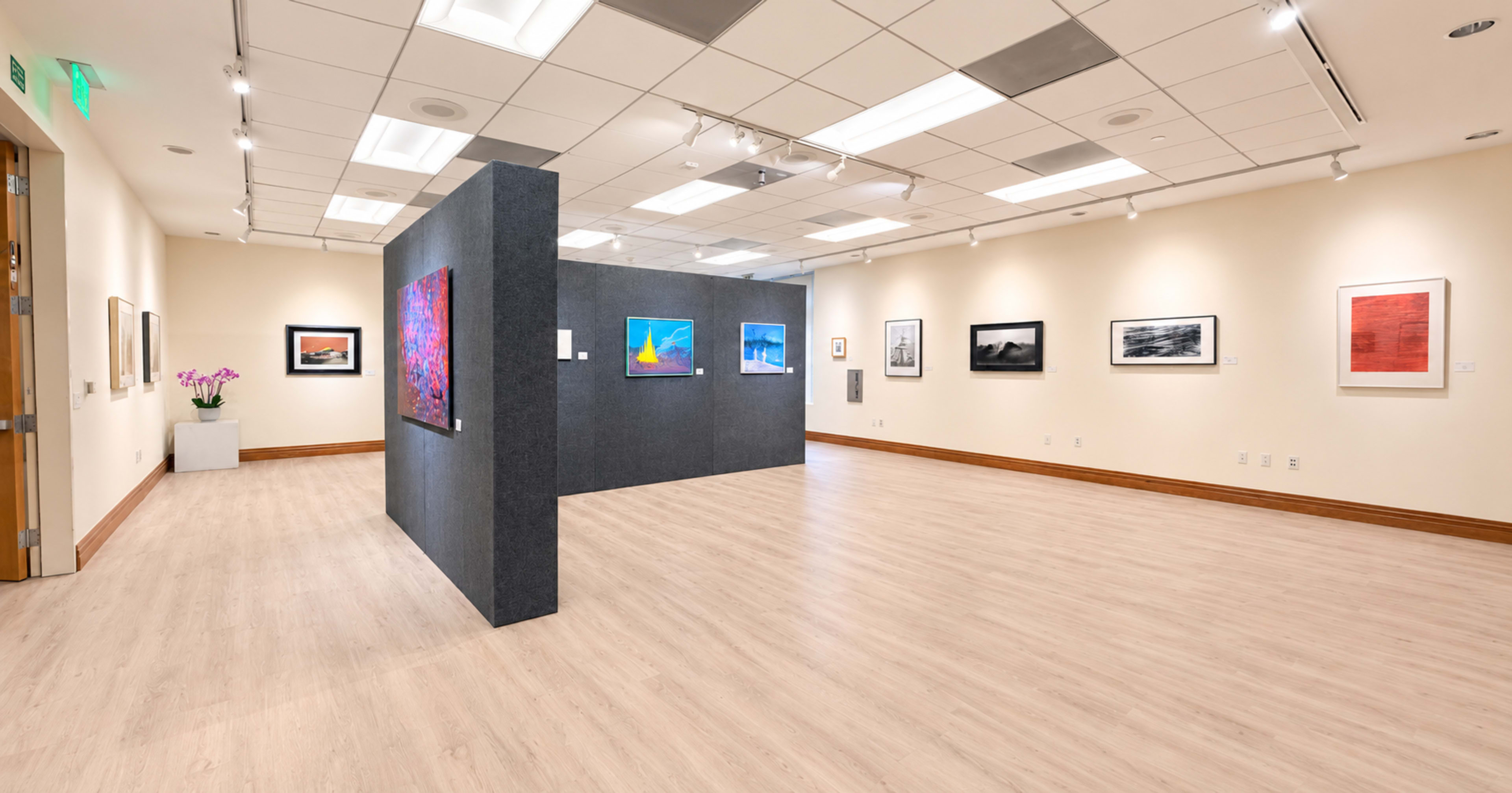 The image shows an art gallery interior featuring several framed artworks displayed on the walls and a central partition with additional pieces.