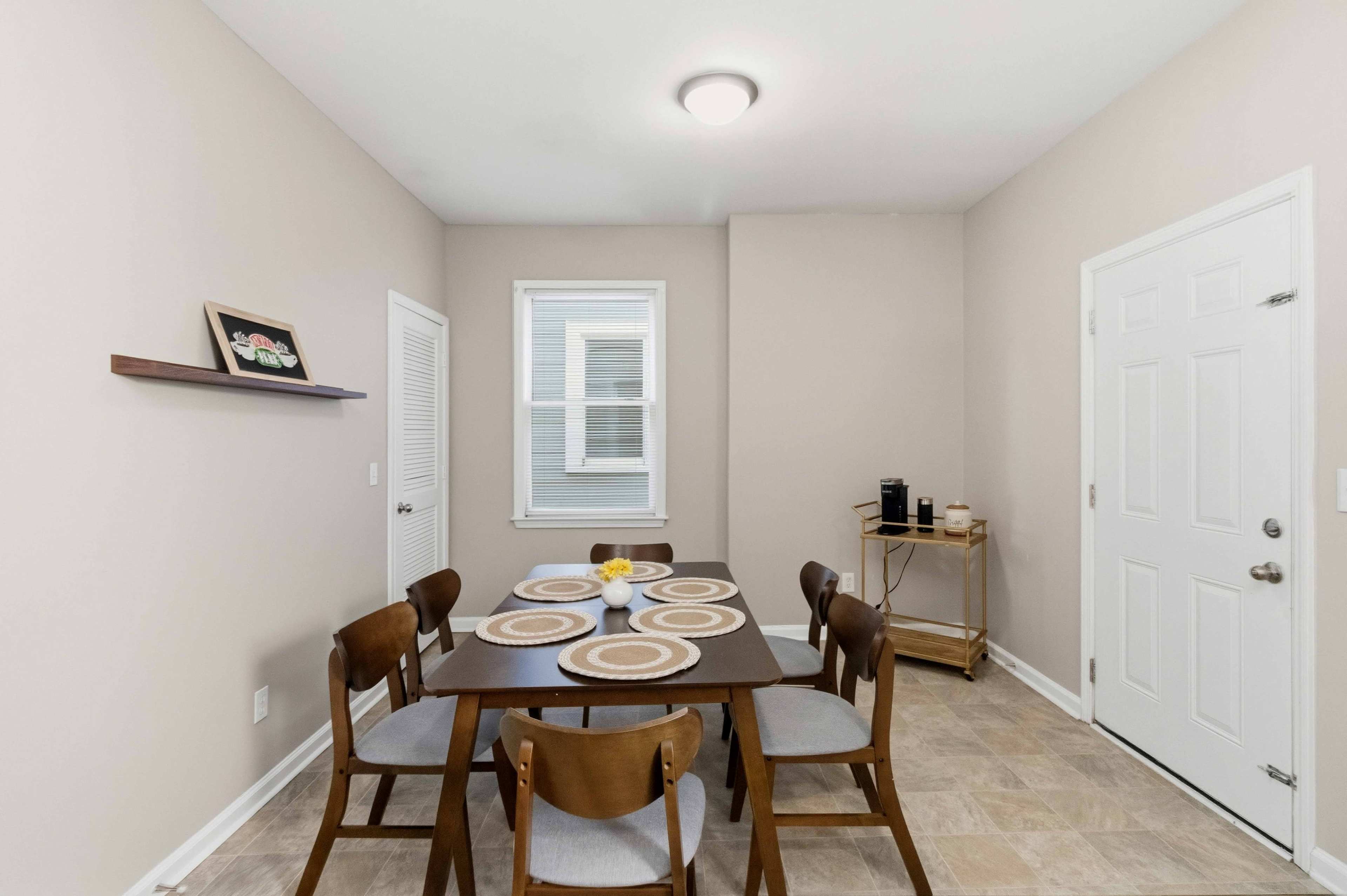 A small kitchen dining area features a wooden table with four chairs, a patterned tablecloth, and a side cart next to a door.