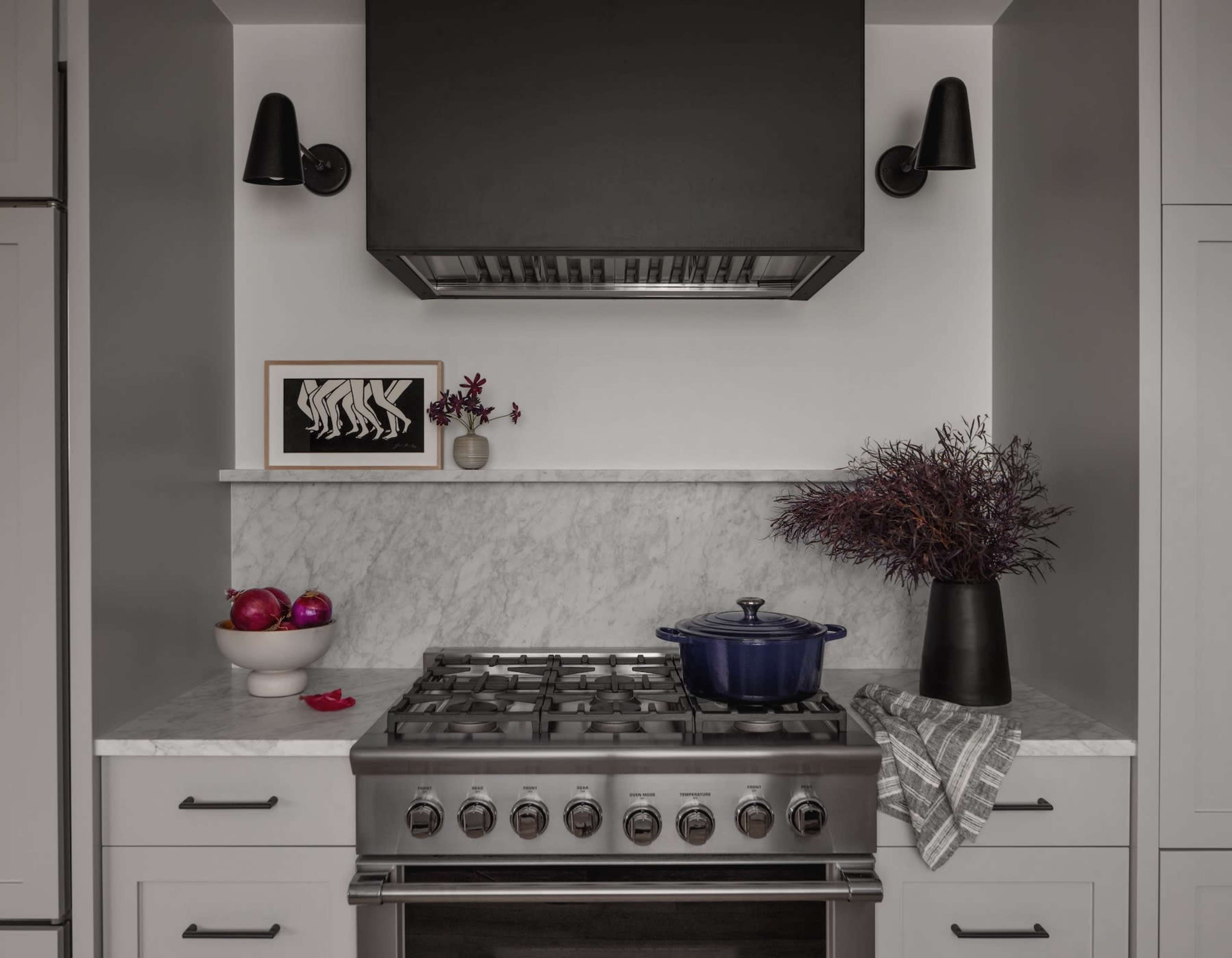 A modern kitchen features a gas stove, a marble countertop with a dark pot, fruits, and decorative items, complemented by wall-mounted lights and a framed artwork.