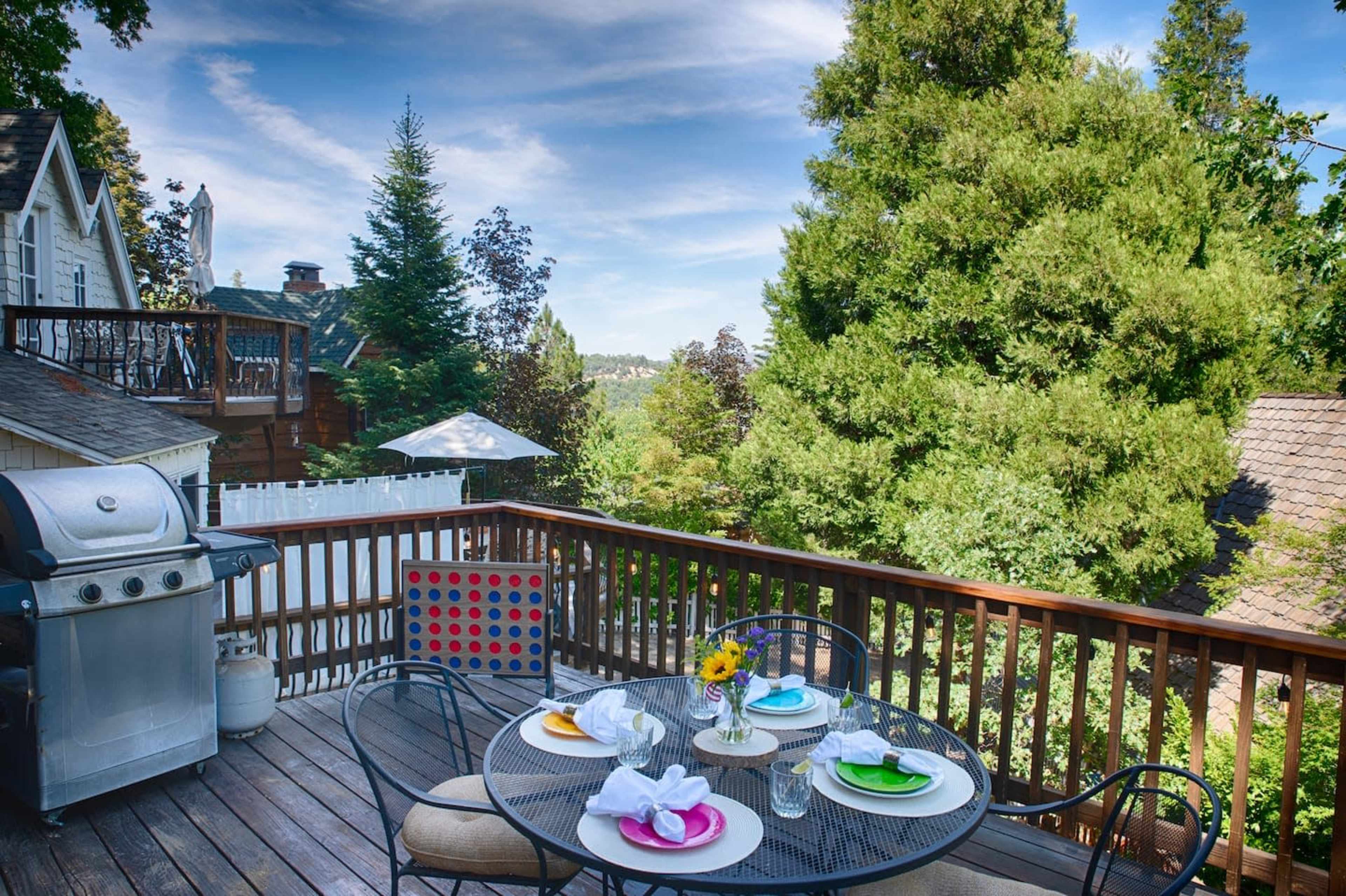 The image shows a wooden deck with a table set for dining, featuring colorful plates and a flower centerpiece, alongside a barbecue grill and a view of trees in the background.