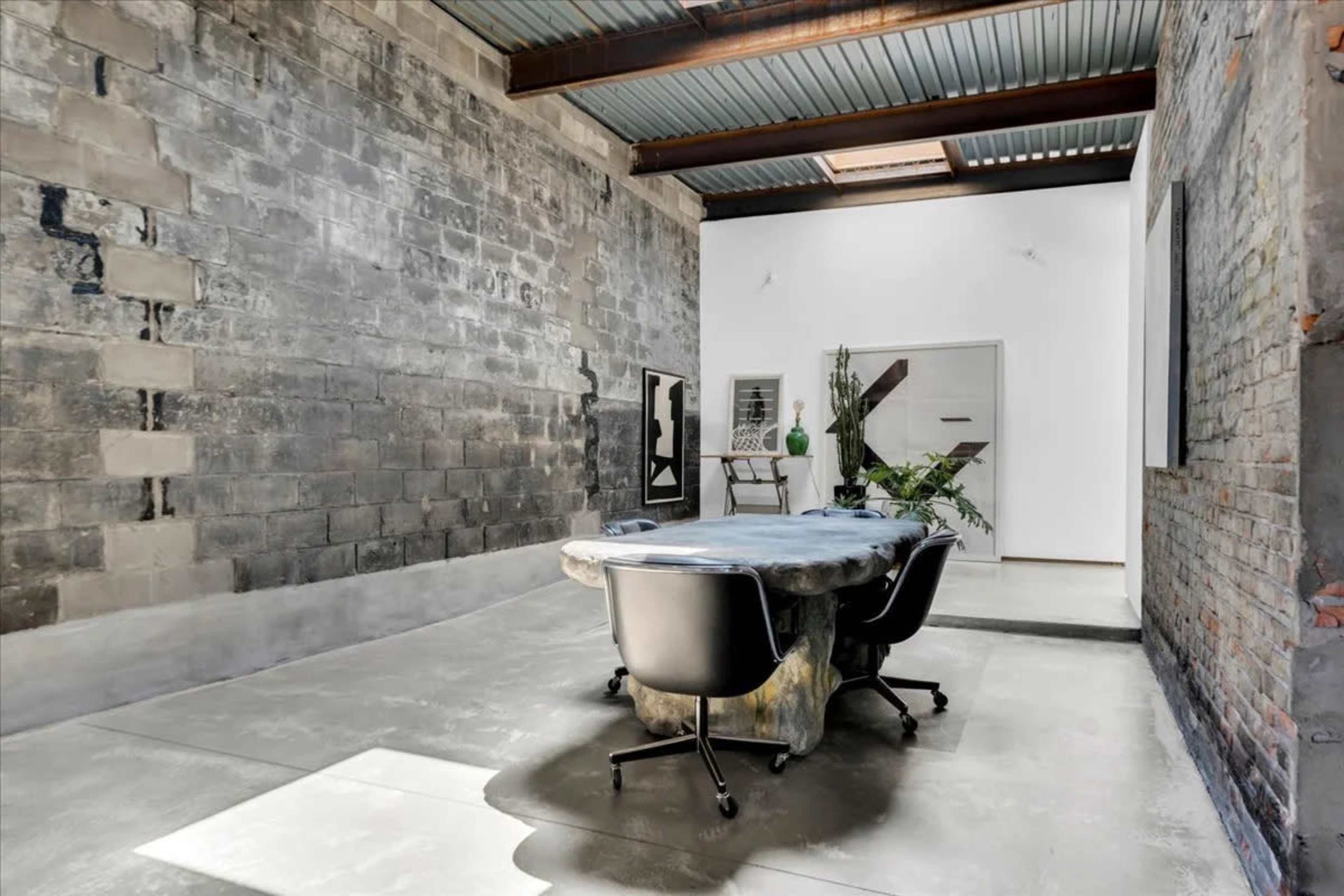 A minimalist workspace features a large, irregular table surrounded by black chairs, set against a raw, exposed brick and concrete wall.