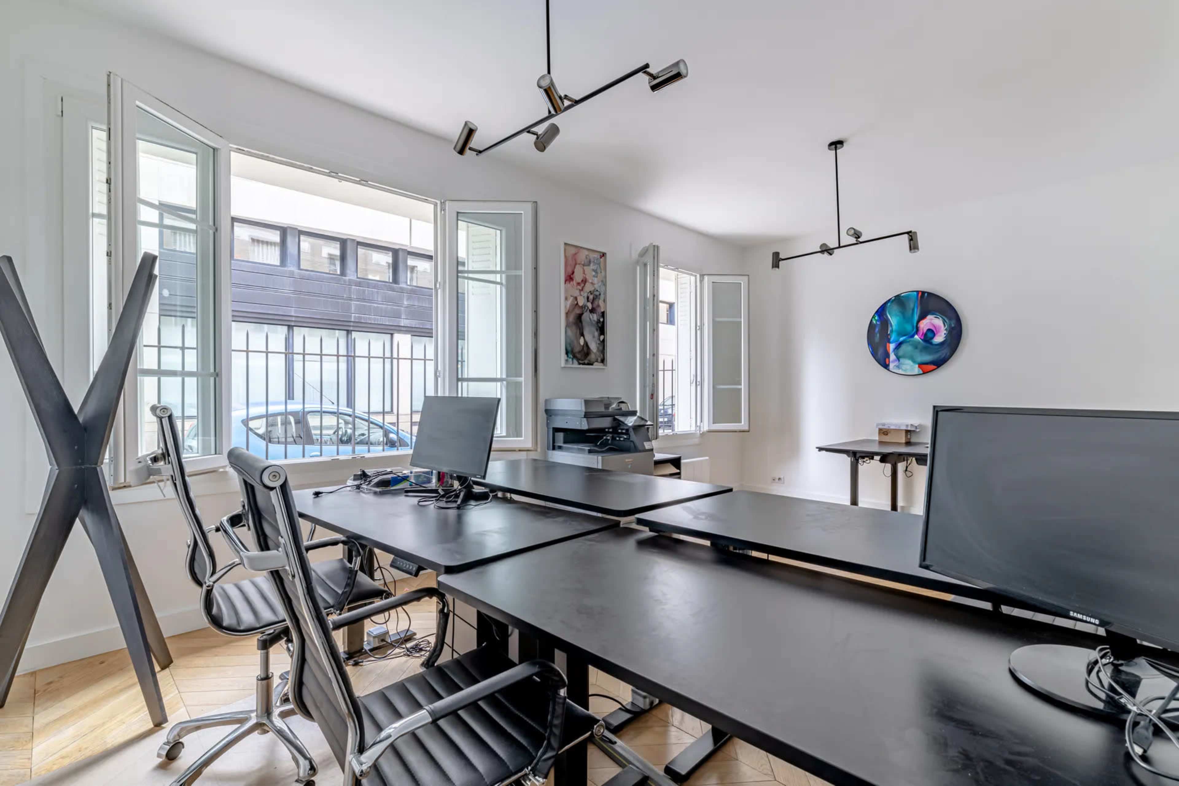A minimalist office space features black desks, ergonomic chairs, and large windows allowing natural light, with a printer and artwork on the walls.