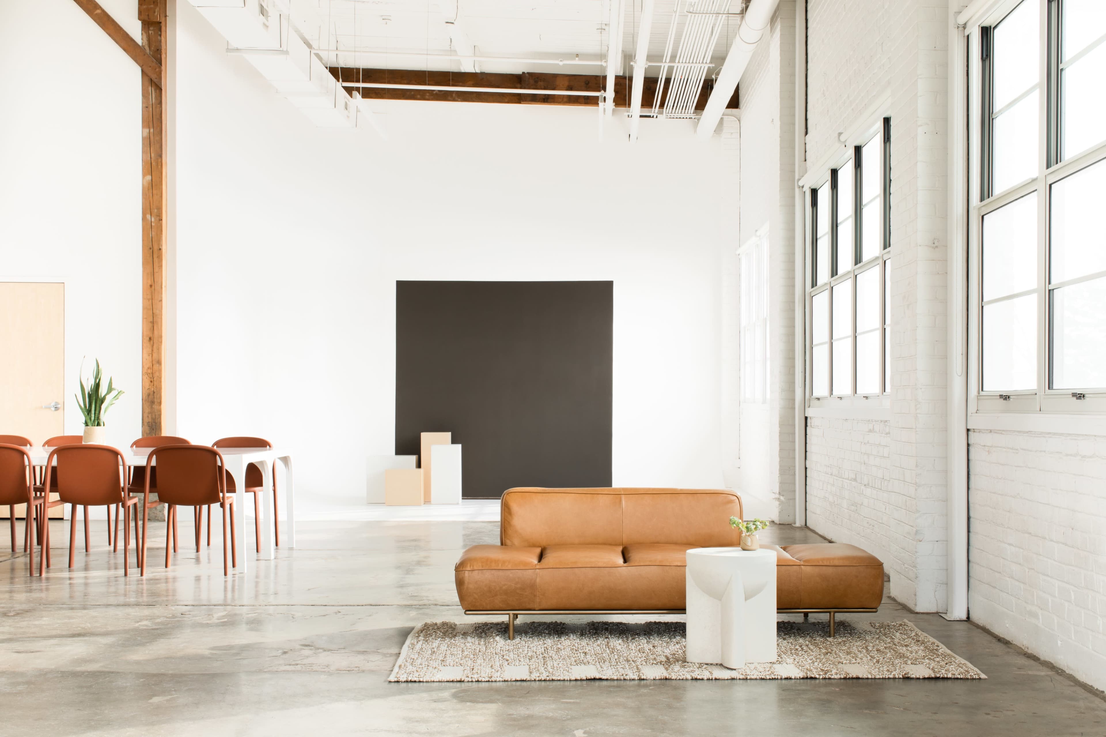 A leather sofa sits on a rug in a spacious room with large windows, a black wall, and a dining area featuring wooden chairs and tables.