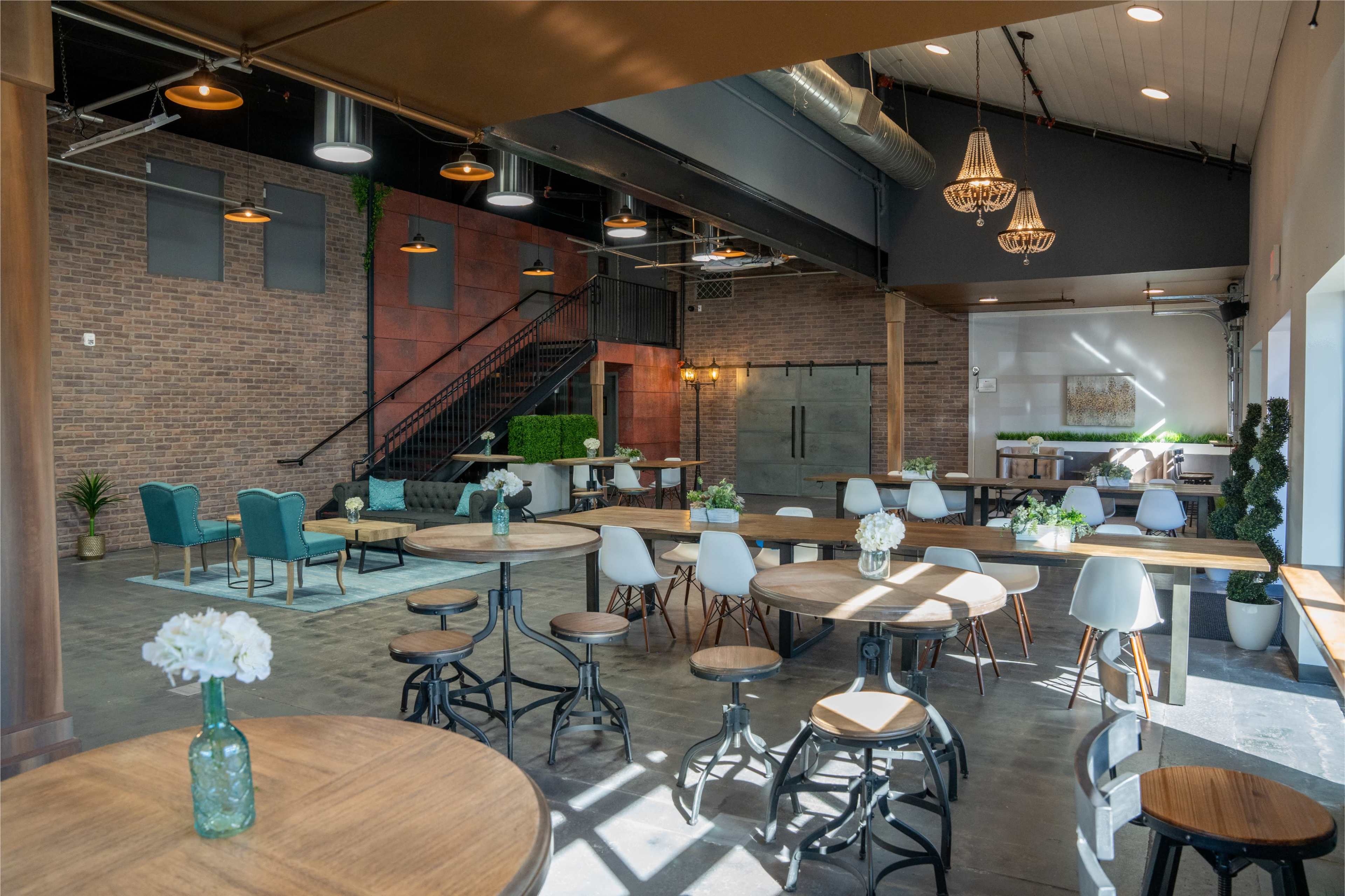 A spacious indoor area with a combination of wooden tables, chairs, and plants, featuring a staircase in the background and large windows that allow natural light to enter.