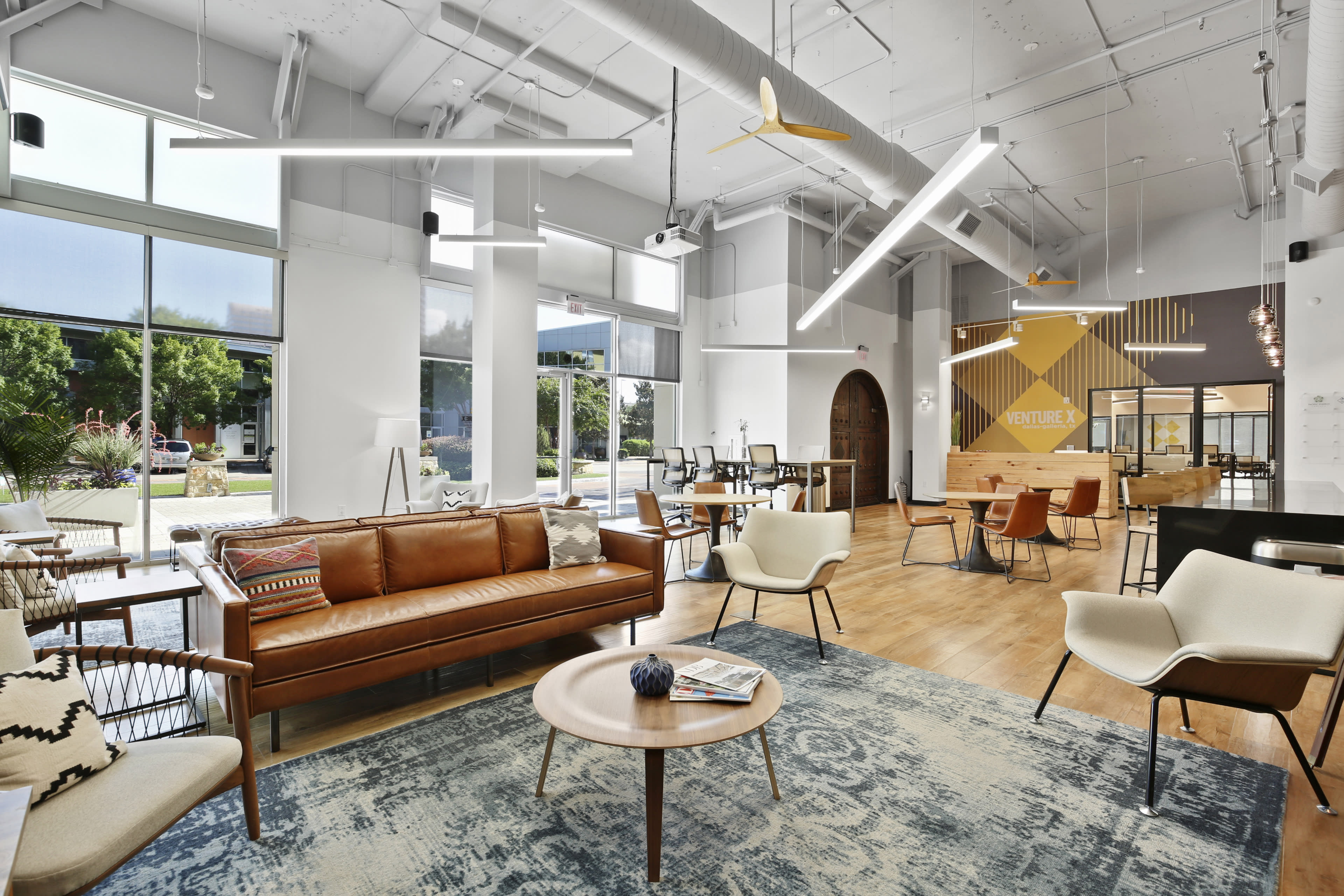 The image shows a modern, shared workspace featuring a spacious seating area with a leather couch, light wooden tables, and large windows that let in natural light.