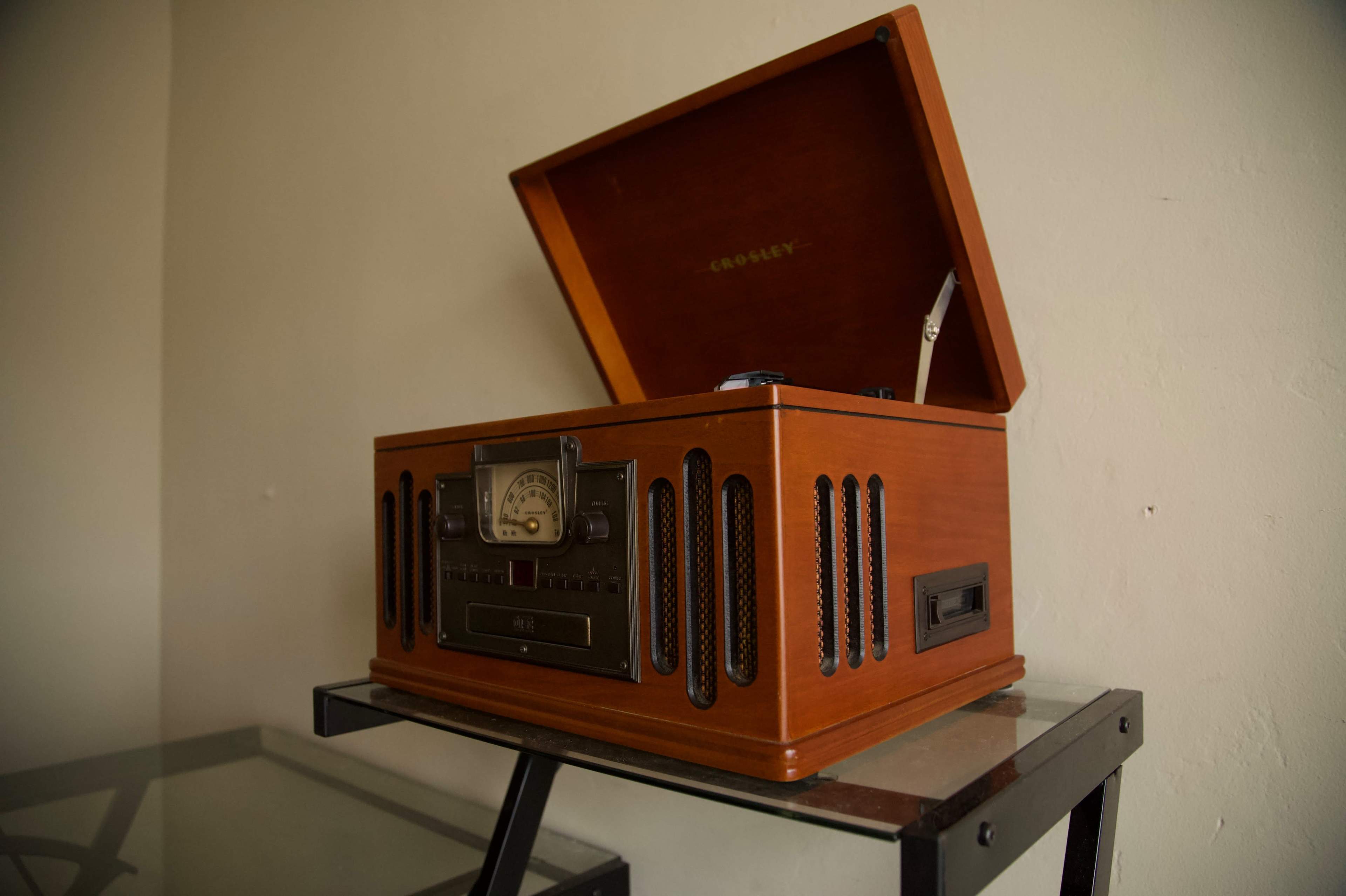 A vintage wooden record player sits on a glass table against a plain wall.