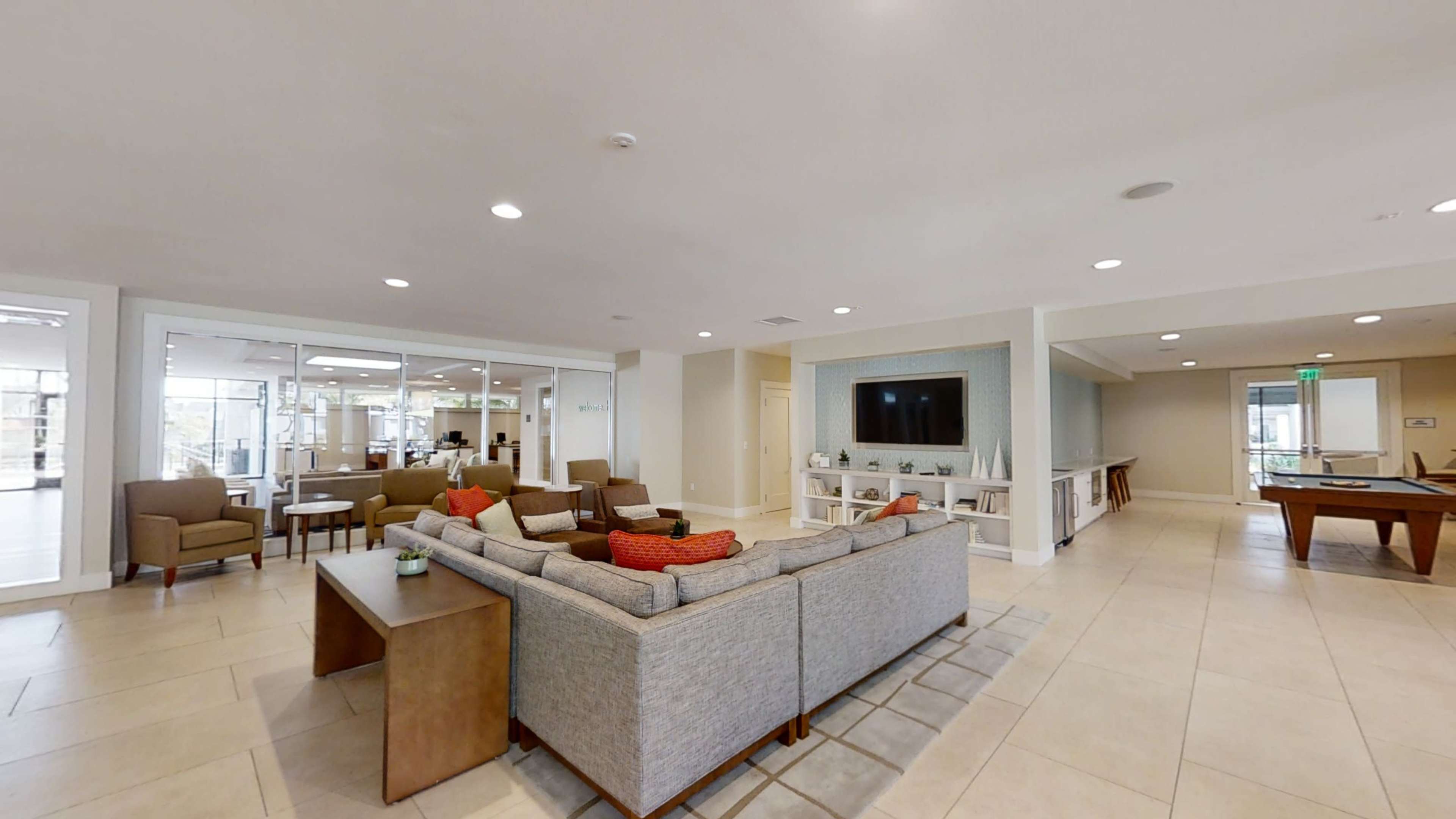 The image shows a modern lounge area with a sectional sofa, chairs, and a large television in an airy common space.