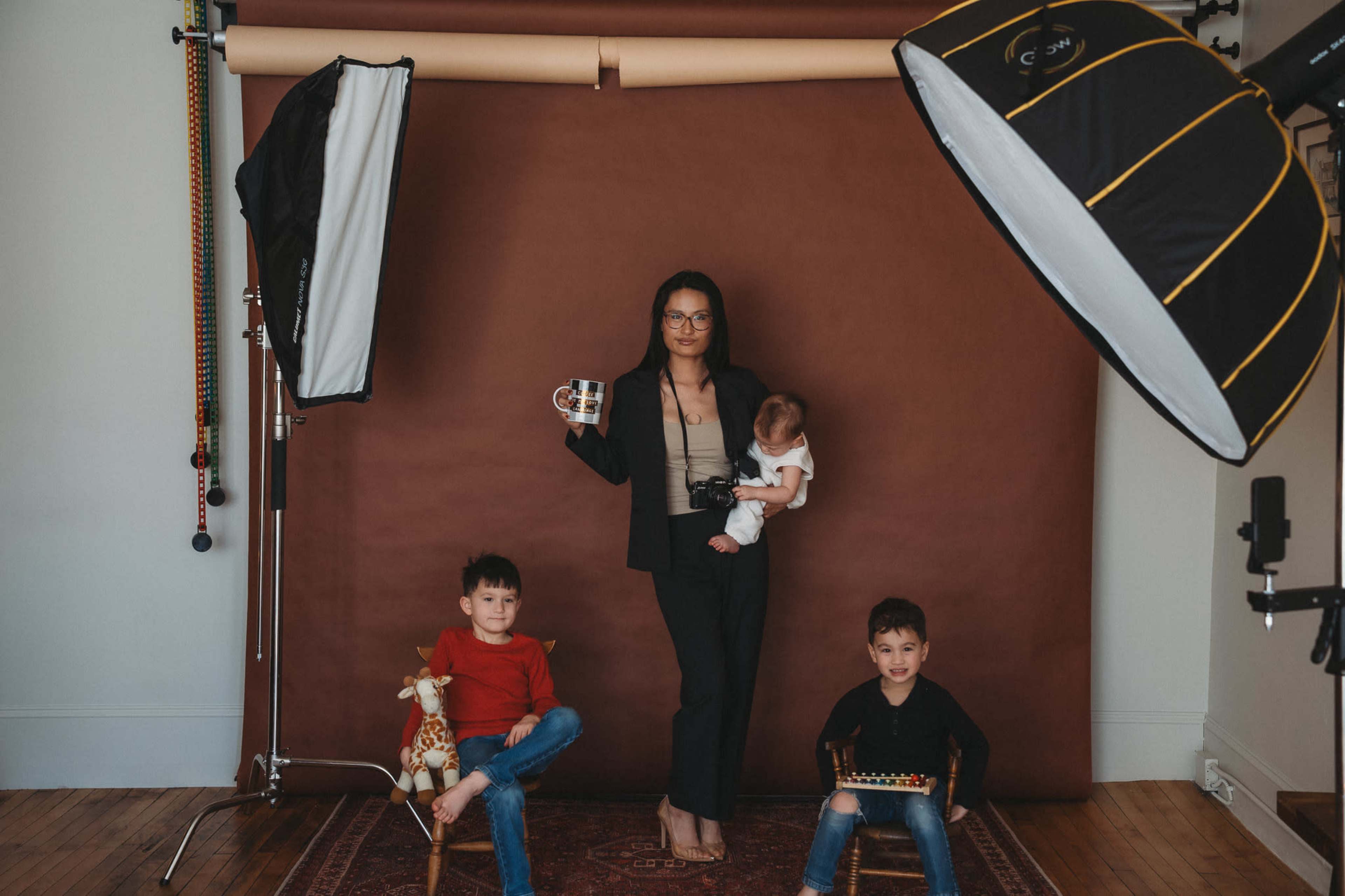 A woman holds a baby and a mug while two young boys sit on chairs beside her in a studio setting.