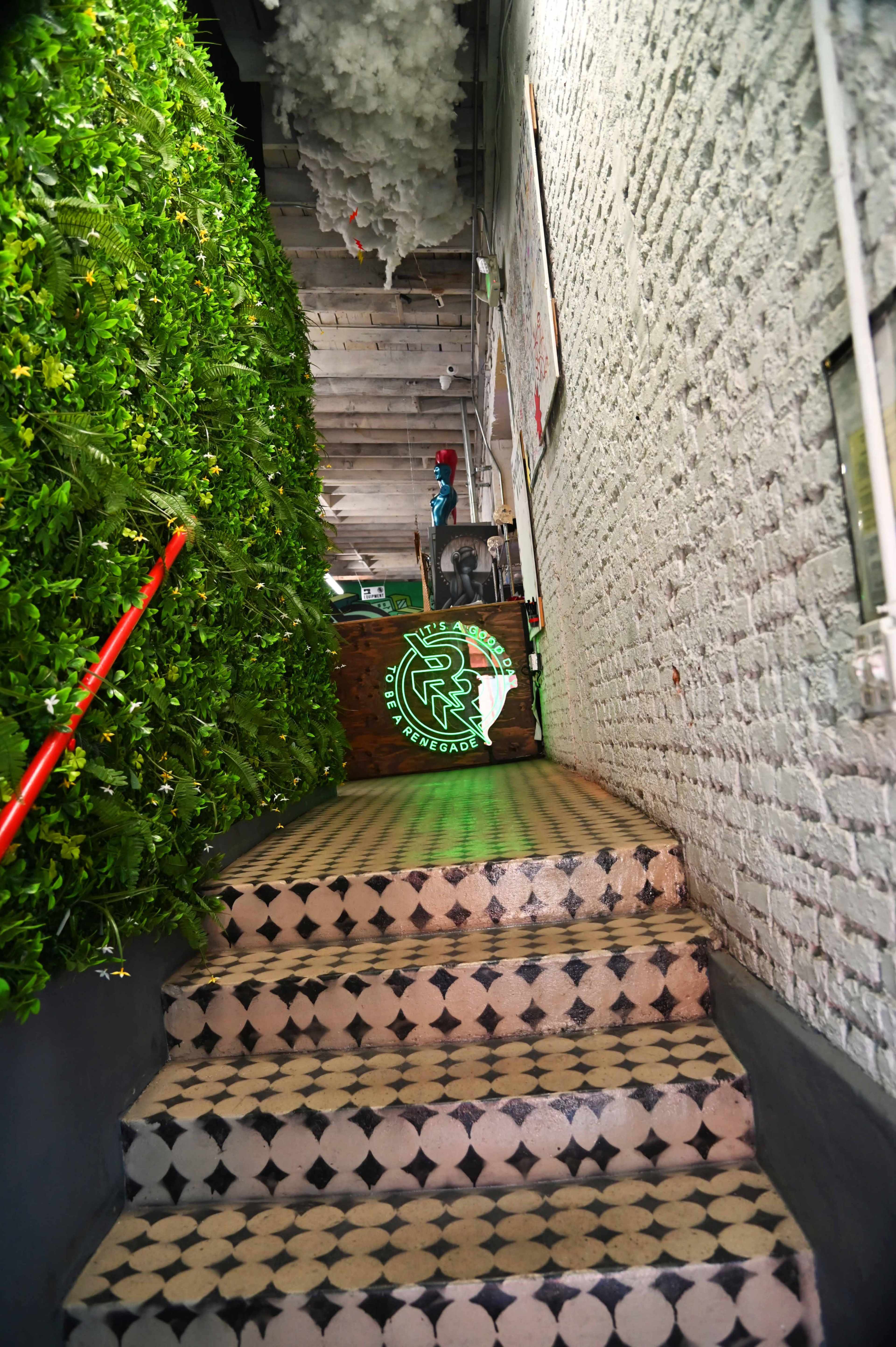 The image shows a staircase lined with greenery on one side, leading up to a decorative wall with a logo at the top.