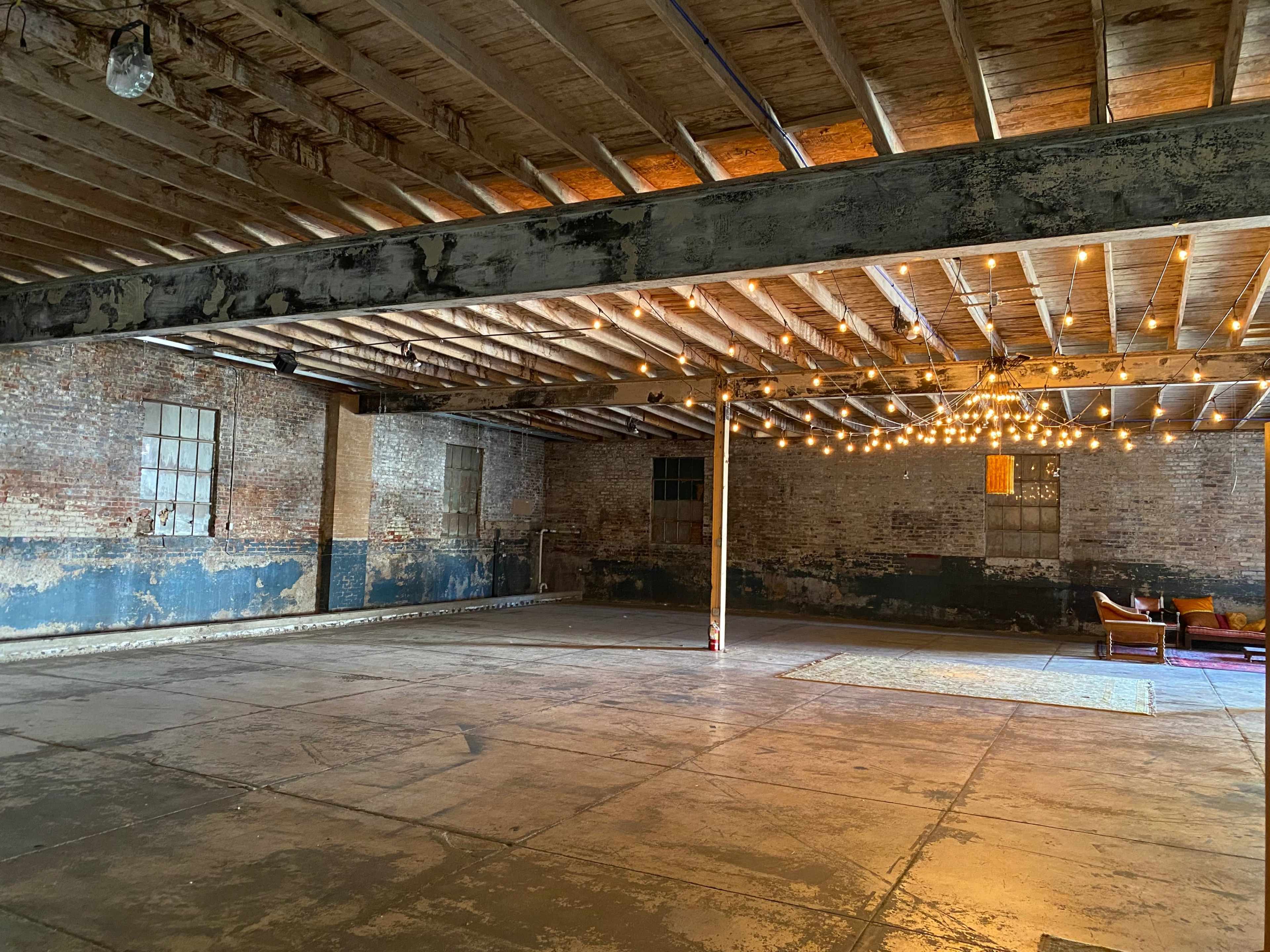 The image shows a spacious, industrial loft with exposed wooden beams, brick walls, and a string light chandelier hanging from the ceiling.