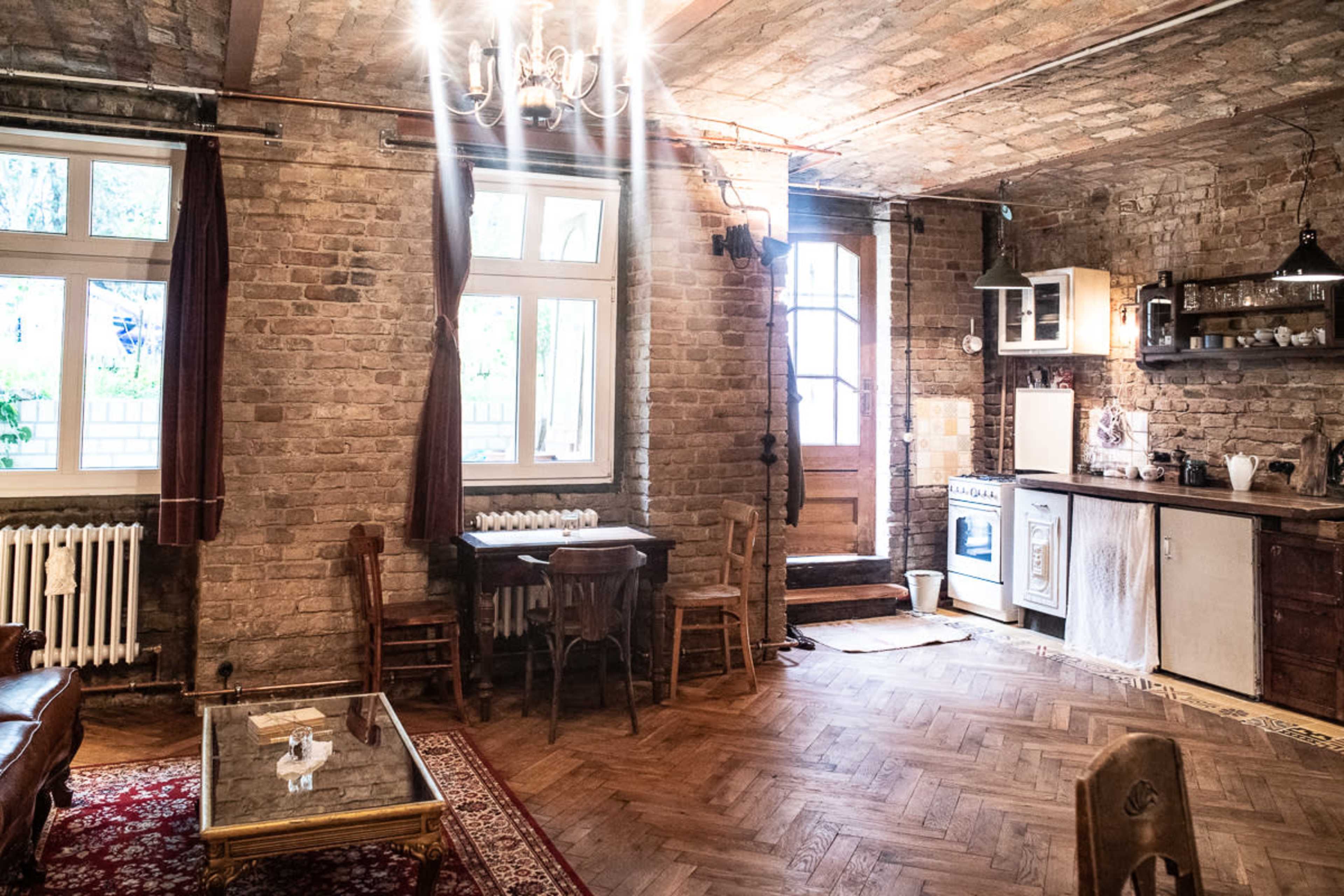 A rustic room with exposed brick walls, a wooden dining table, and a kitchenette, illuminated by a chandelier.