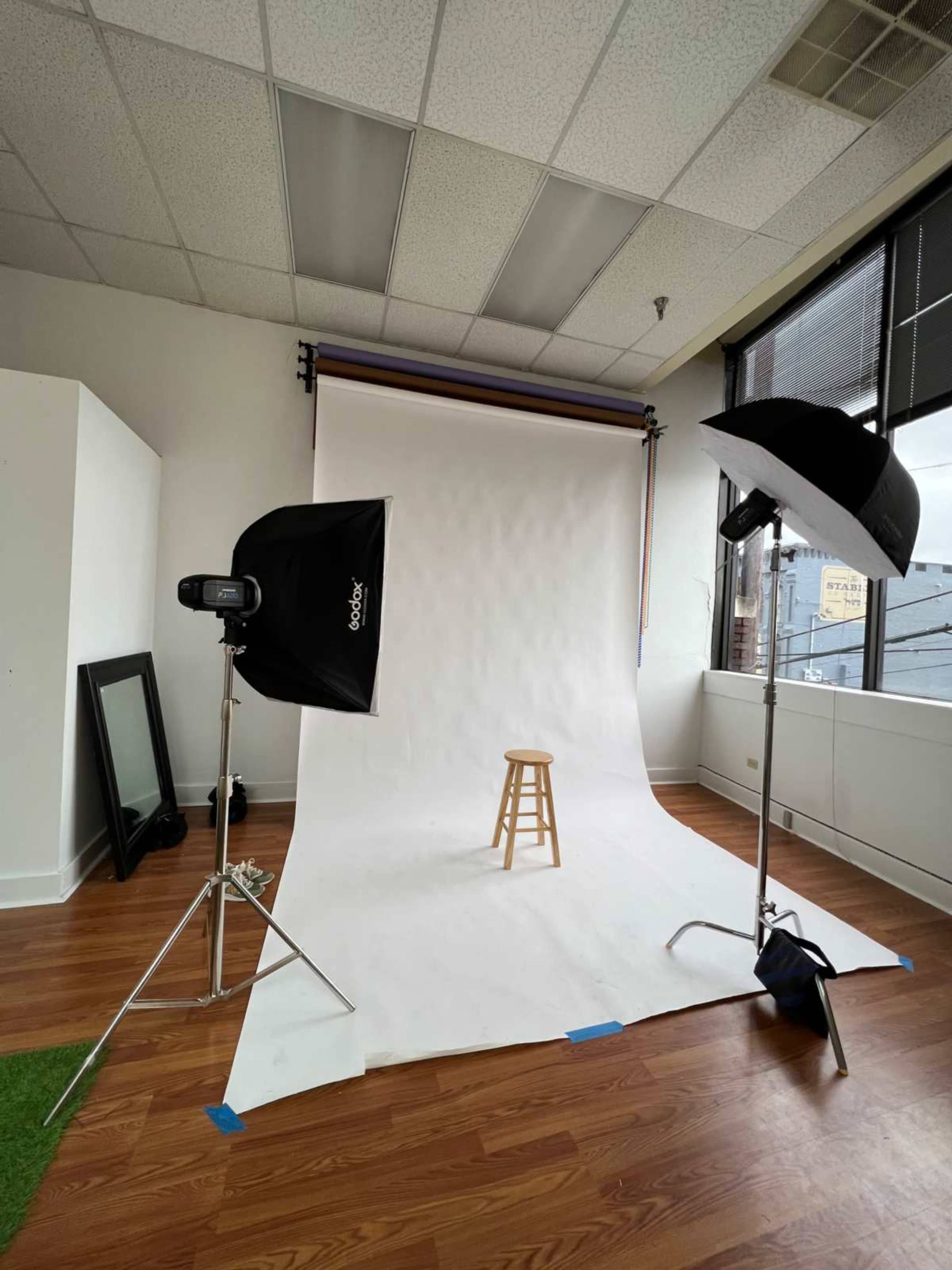 The image shows a photography studio with a blank white backdrop, a wooden stool in the center, and lighting equipment positioned on either side.