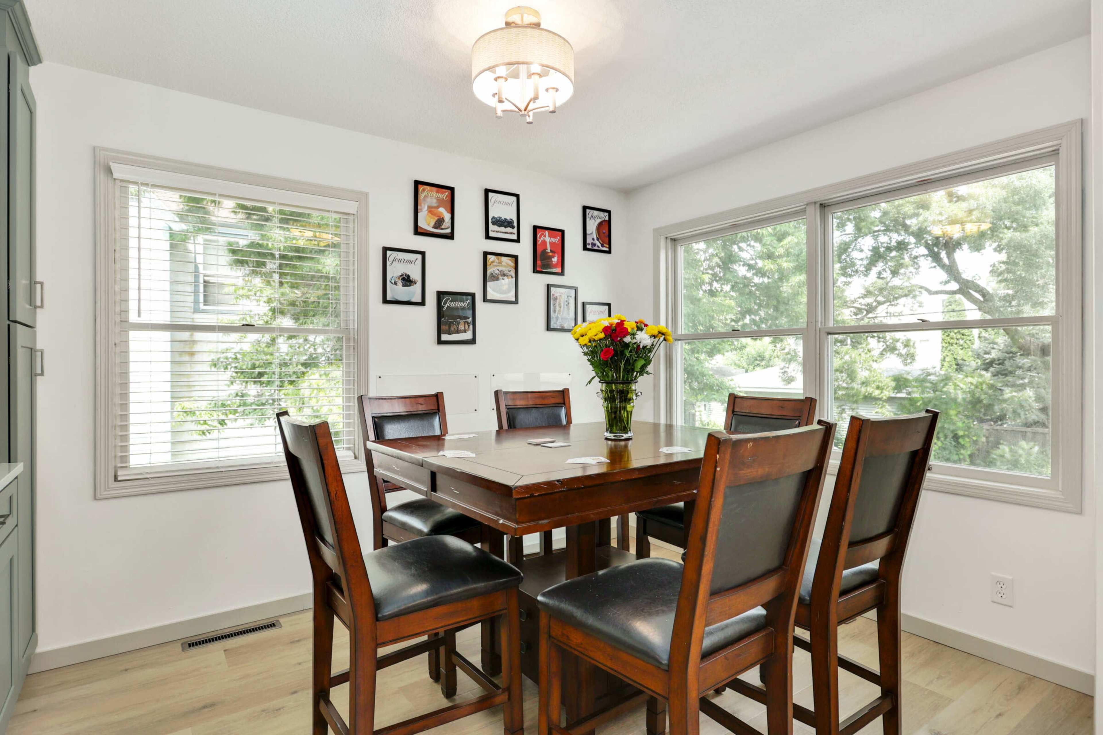 A dining area features a wooden table surrounded by eight chairs, with framed pictures on the walls and a bouquet of flowers on the table.