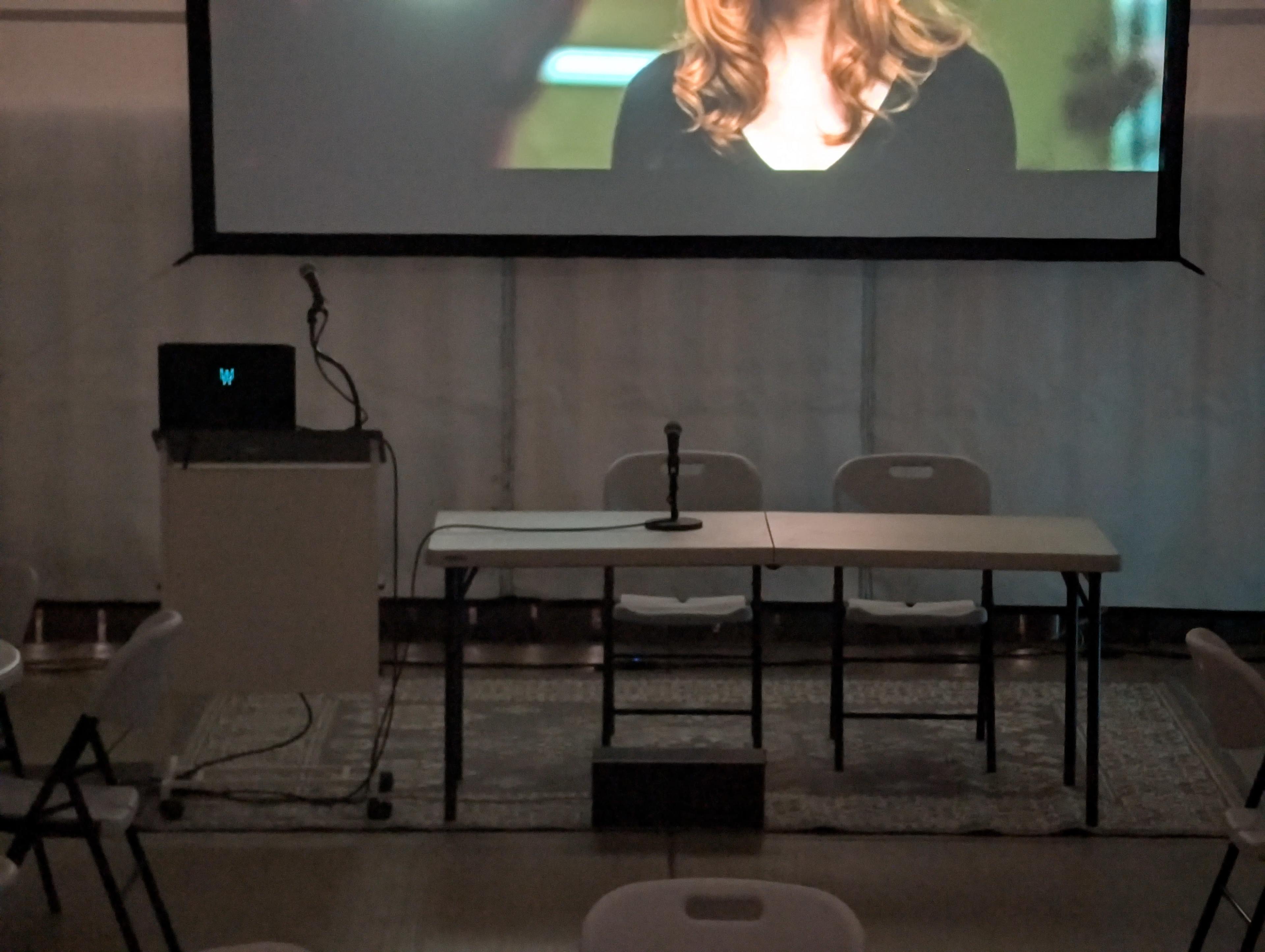 A microphone stands on a table in front of a projected screen displaying a person's face, with empty chairs arranged behind.