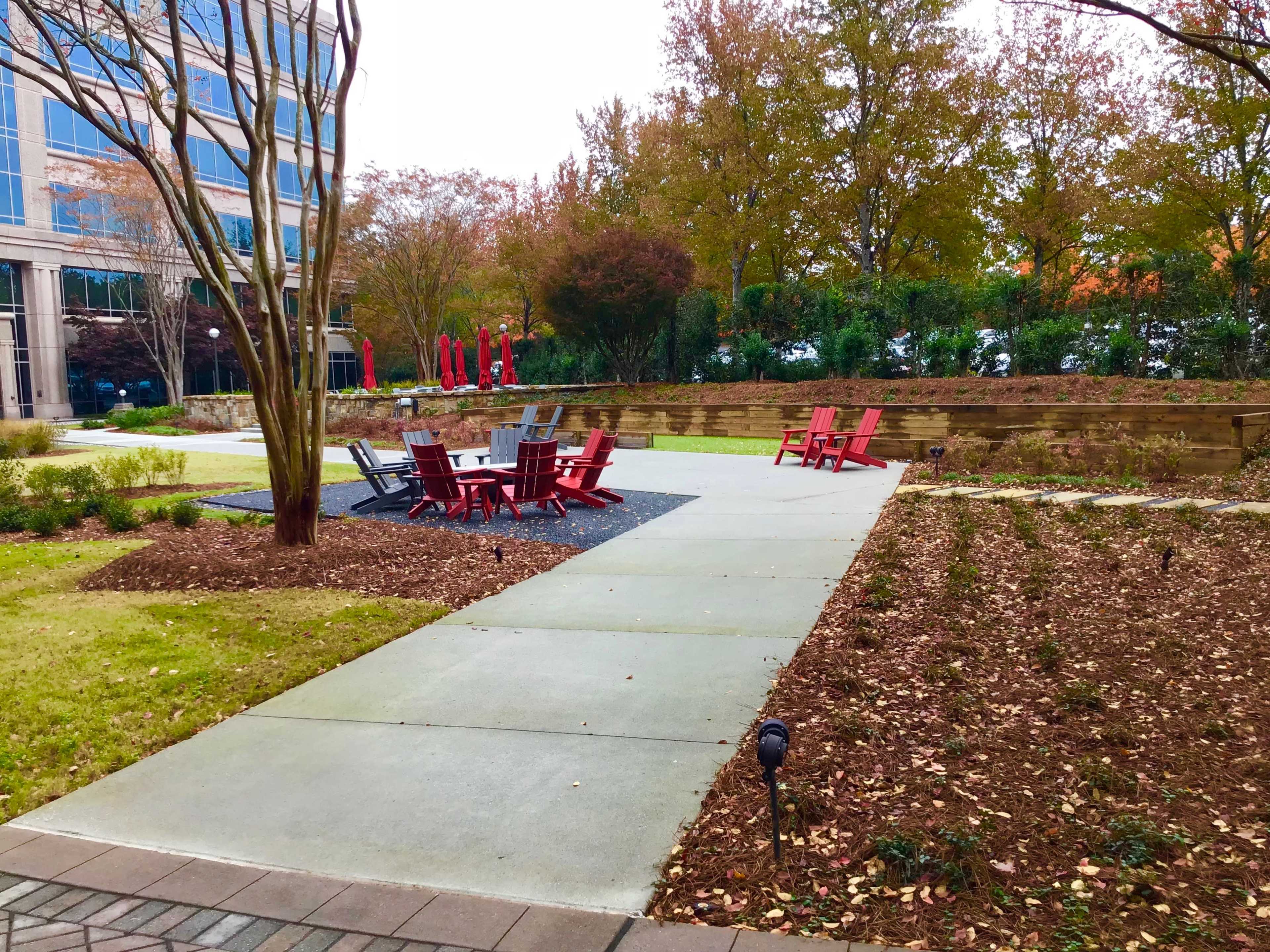 The image shows a landscaped outdoor area with a pathway, seating arrangements made of red chairs, and patches of grass and mulch.