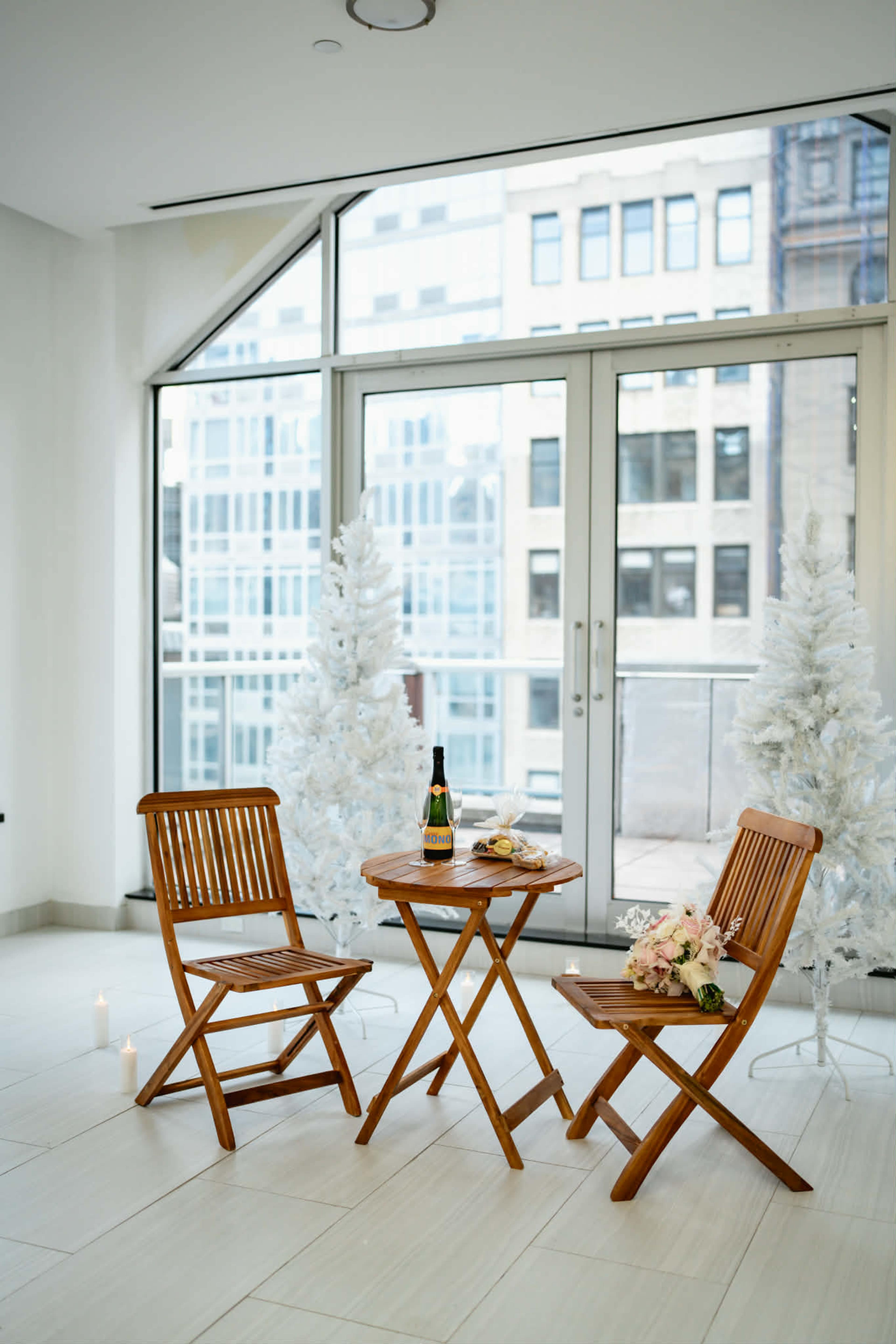 A small wooden table is set between two folding chairs, with a bottle of champagne, glasses, and a bouquet of flowers, against a backdrop of large windows and white holiday trees.