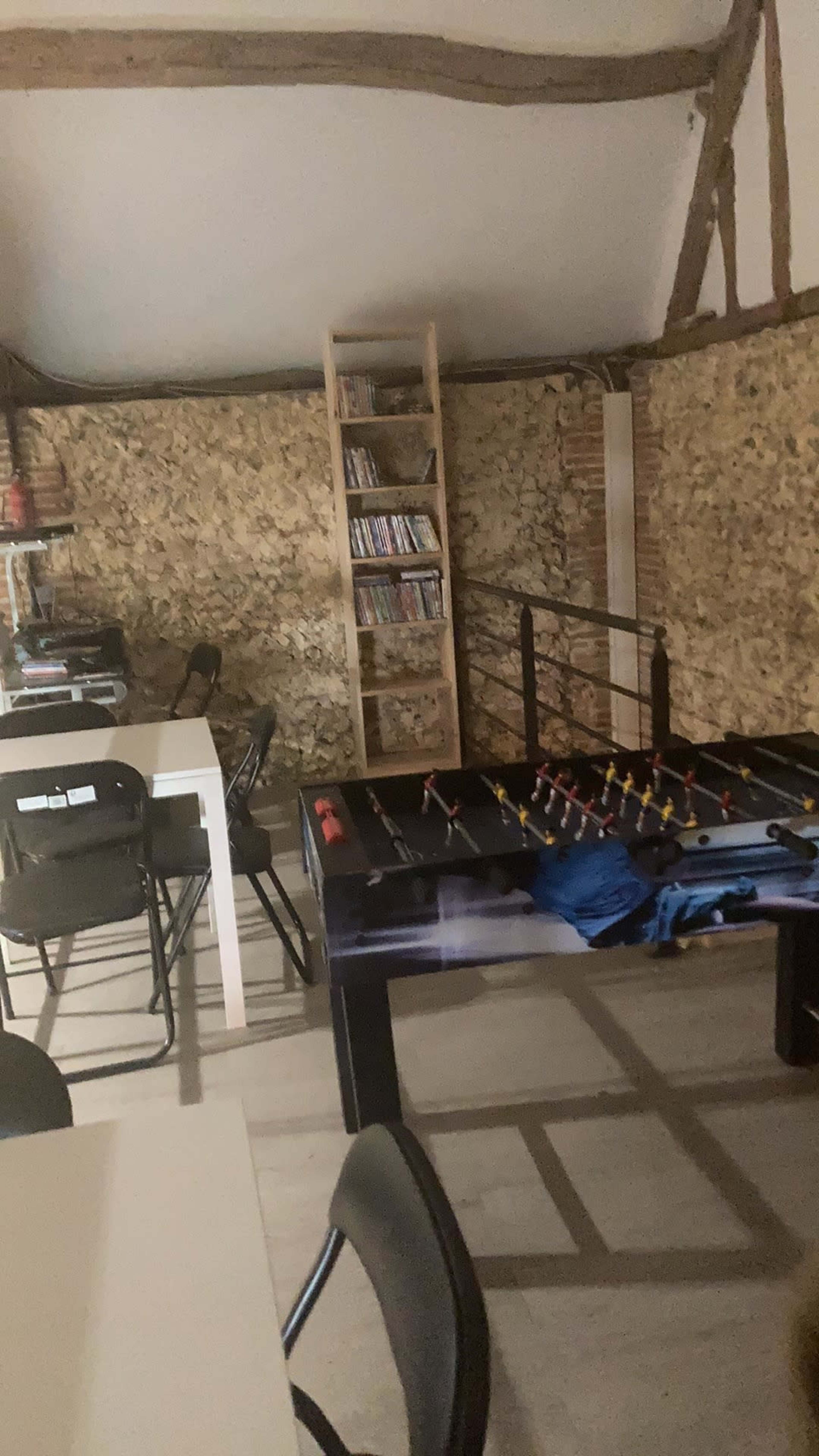 A room with a foosball table, a white table, black chairs, and a bookshelf filled with DVDs against a rustic stone wall.