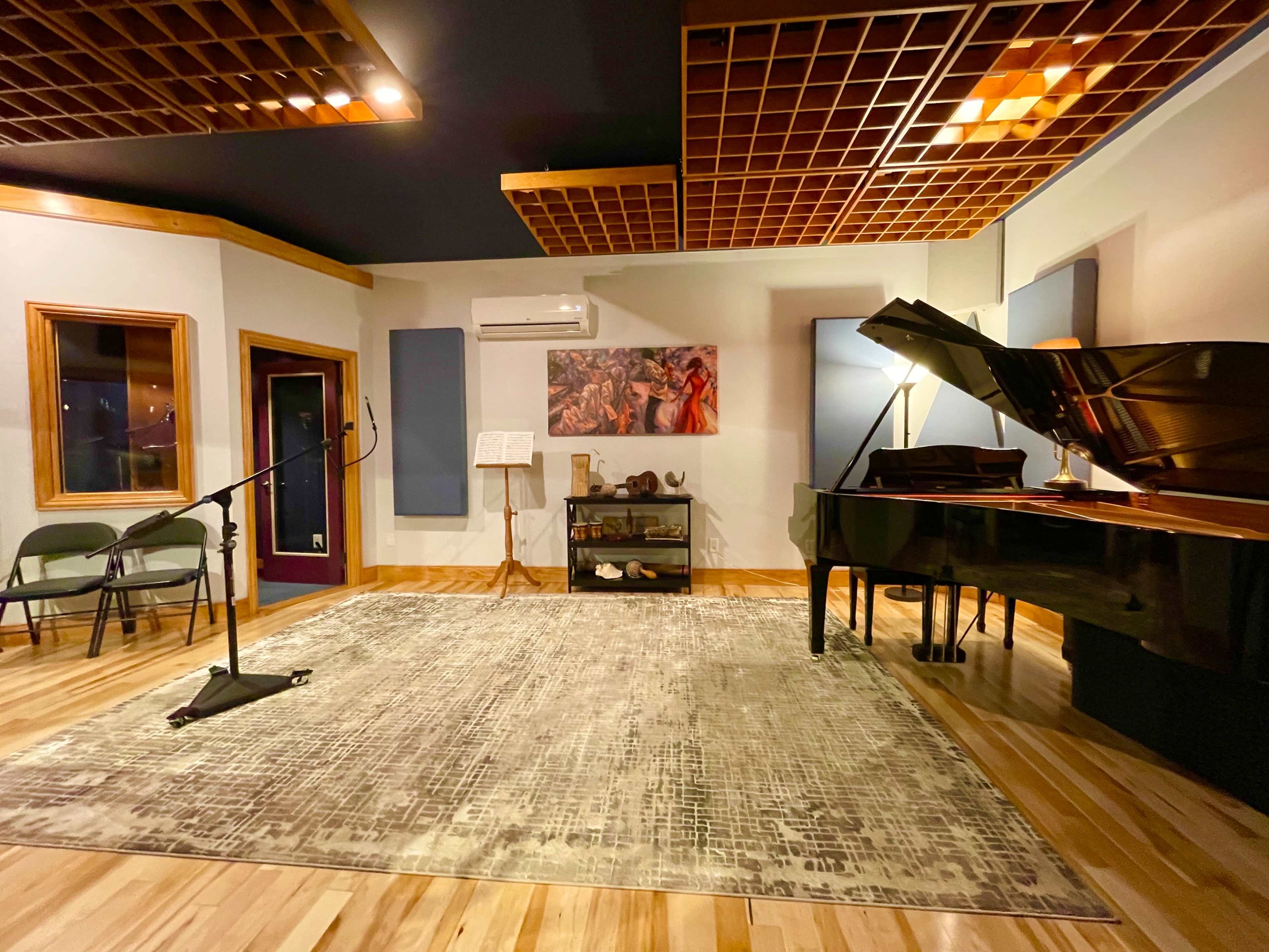 The image shows a music studio with a grand piano, a microphone stand, a sheet music stand, and a large rug on a wooden floor.