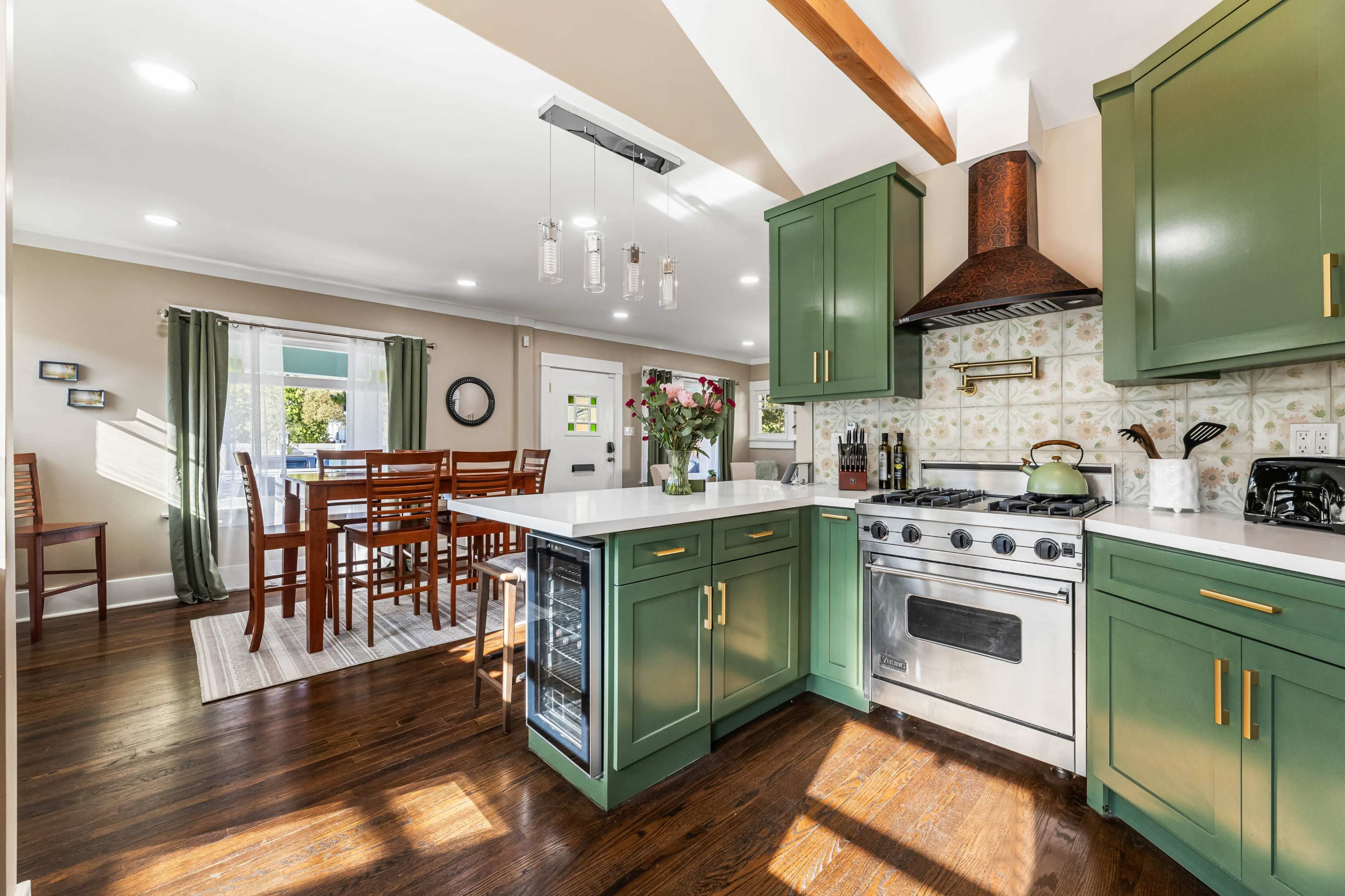 The image shows a modern kitchen featuring green cabinets, a stainless steel stove, and a dining area with wooden chairs adjacent to it.