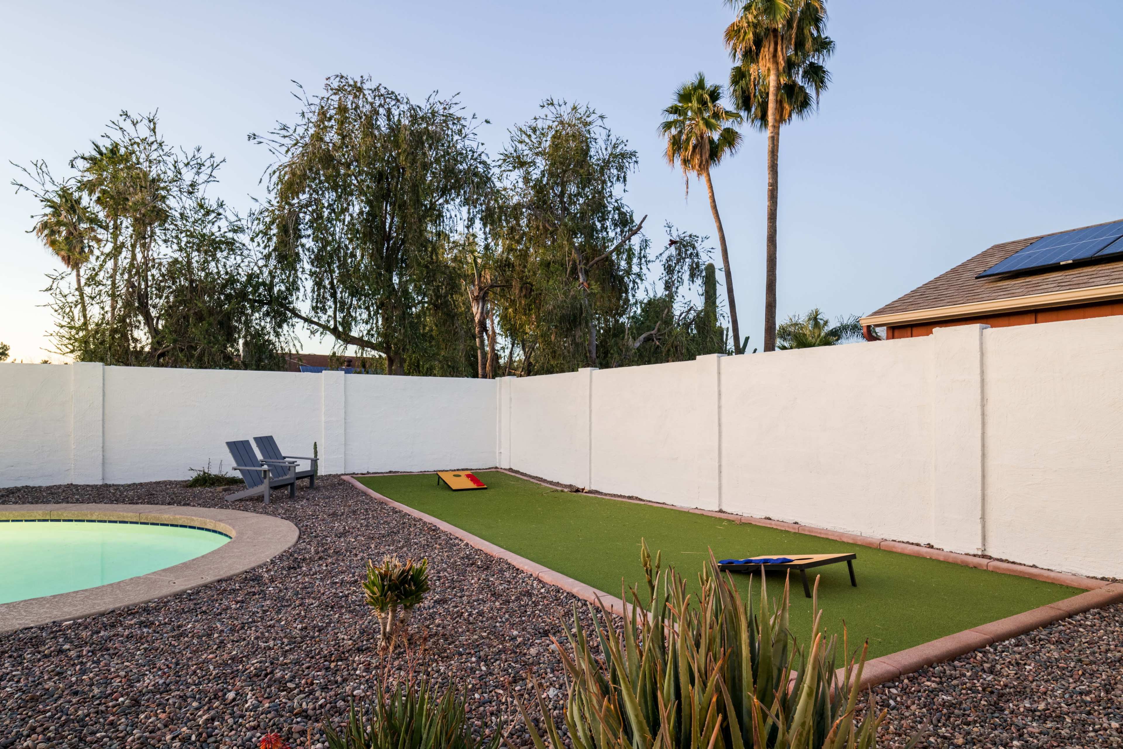 The image shows a backyard with artificial grass, a small lounging area with chairs, a pool, and a white wall surrounding the space.