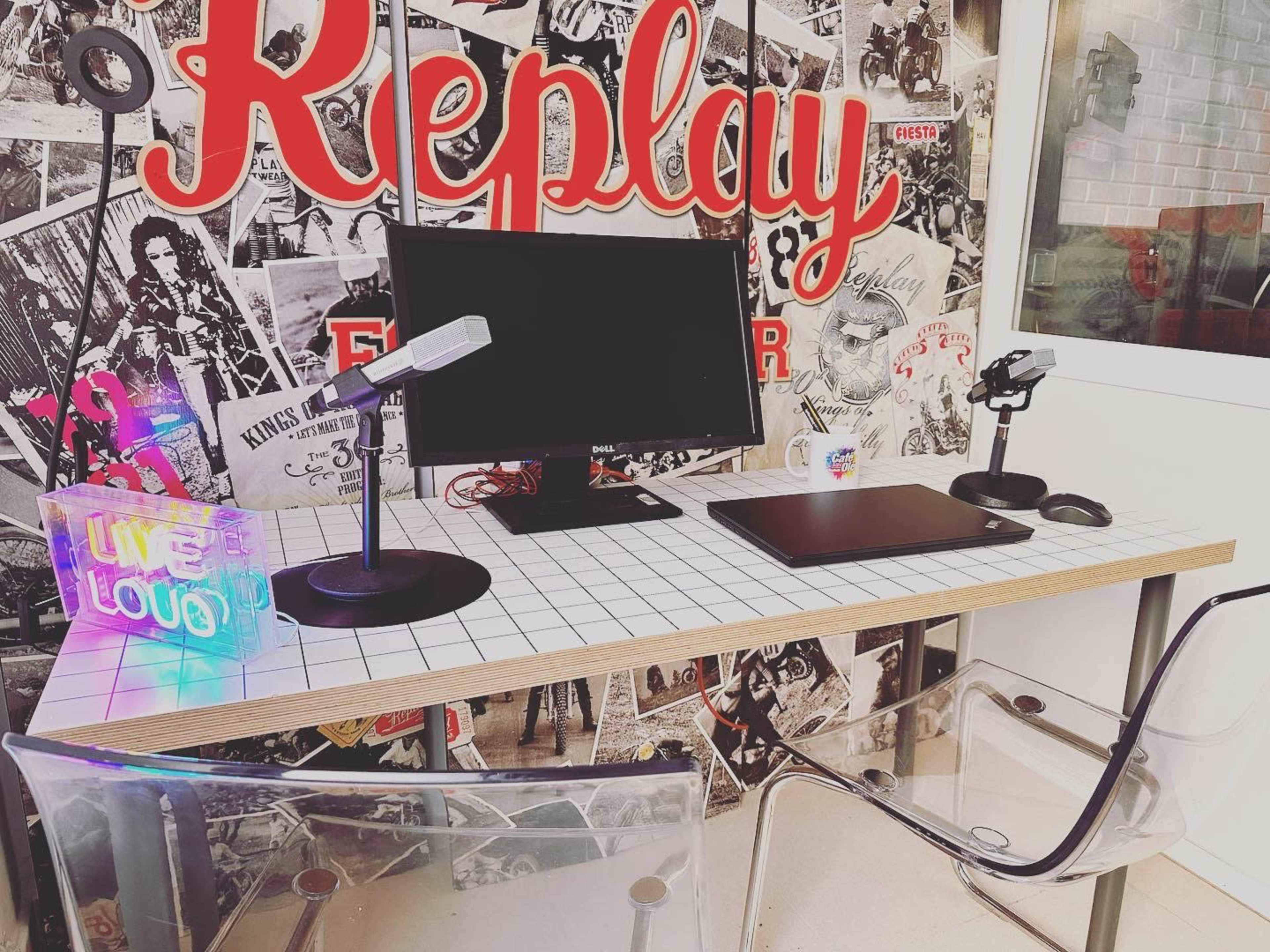 The image shows a modern desk setup featuring a computer monitor, a laptop, a microphone, and colorful decor against a backdrop of black-and-white posters.