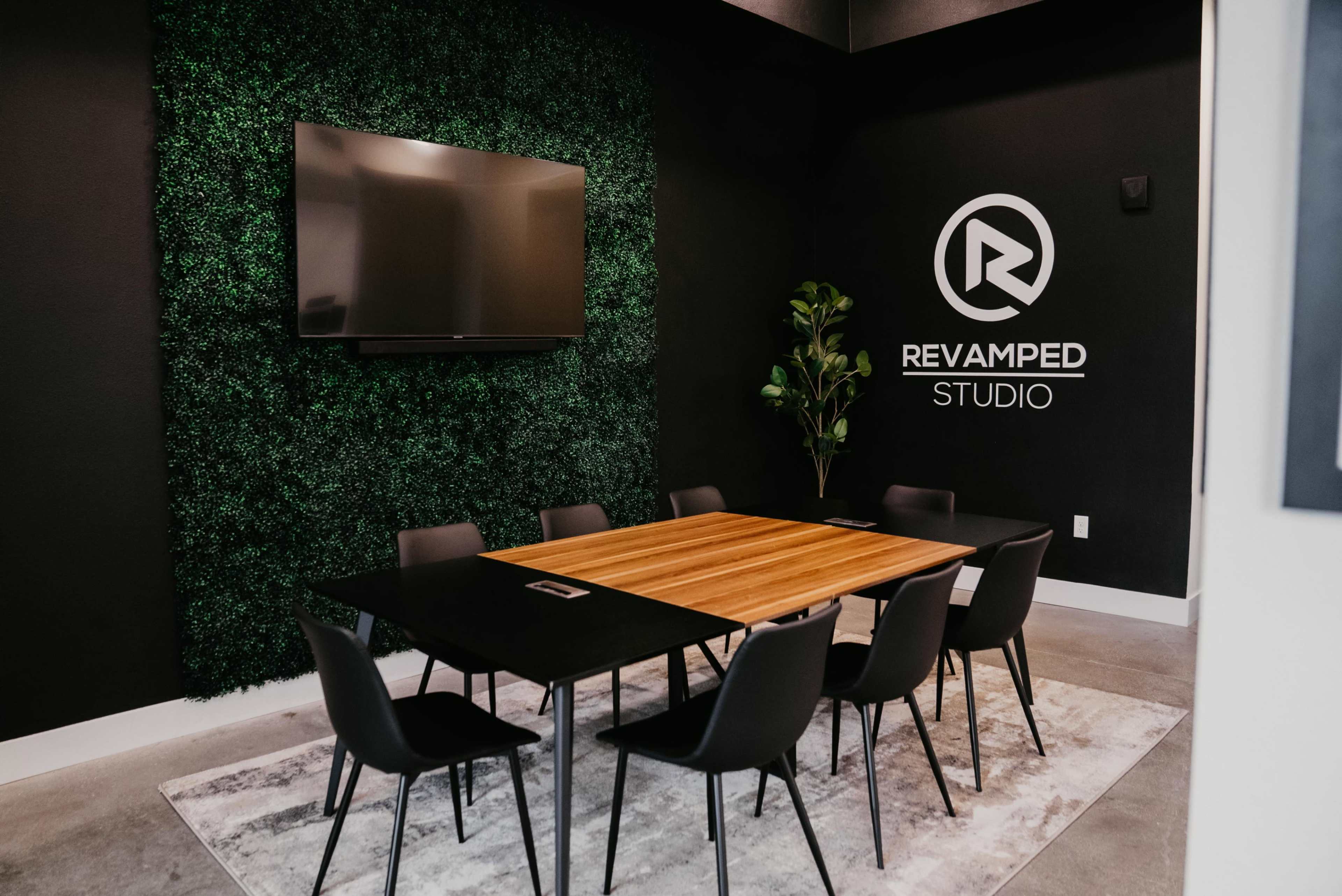 A modern conference room featuring a large wooden table surrounded by black chairs, with a green wall, a television, and a logo for "Revamped Studio."