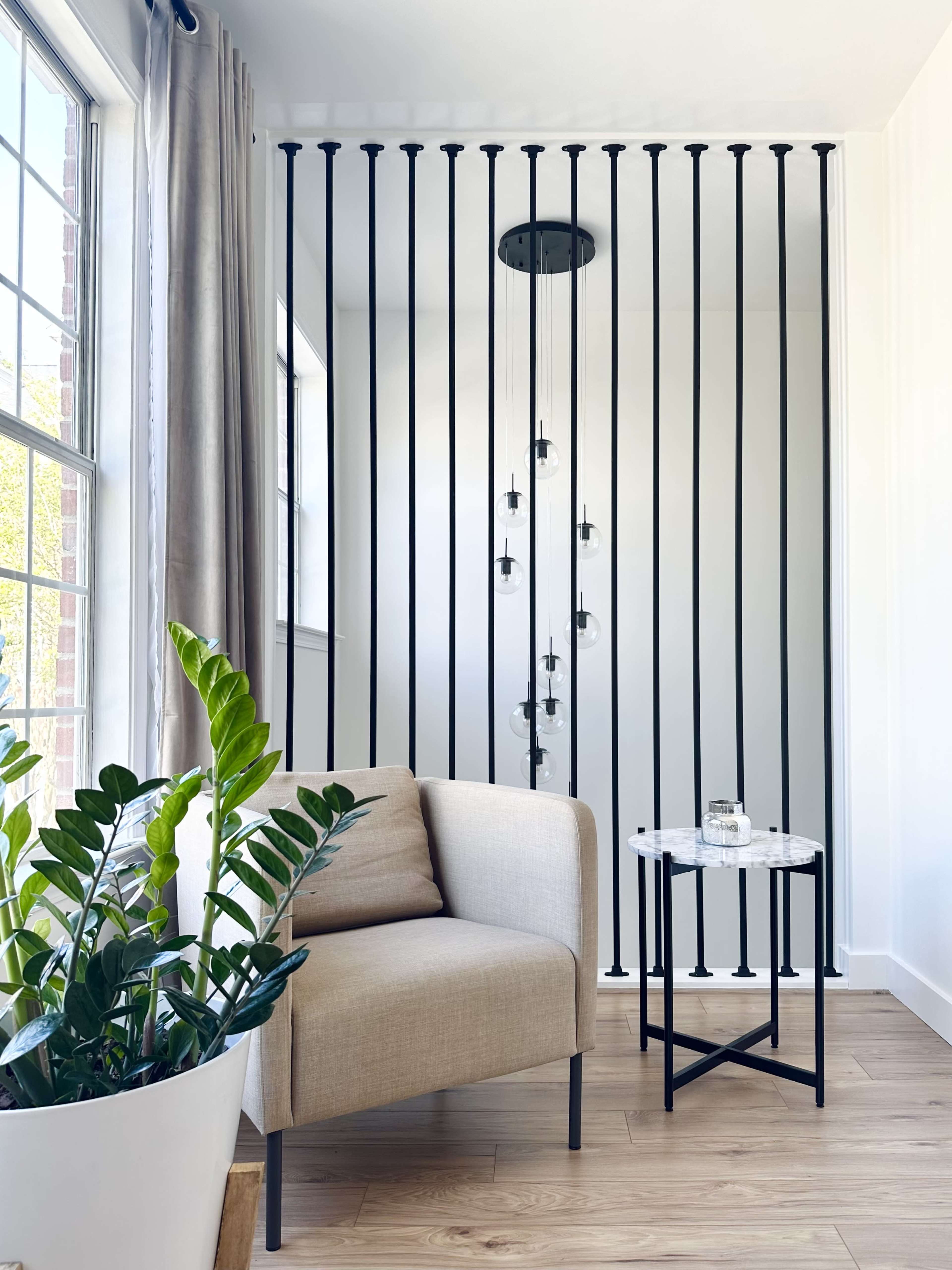 A beige armchair and side table are positioned beside a planter with a plant, separated from a well-lit area by vertical black bars and hanging light fixtures.
