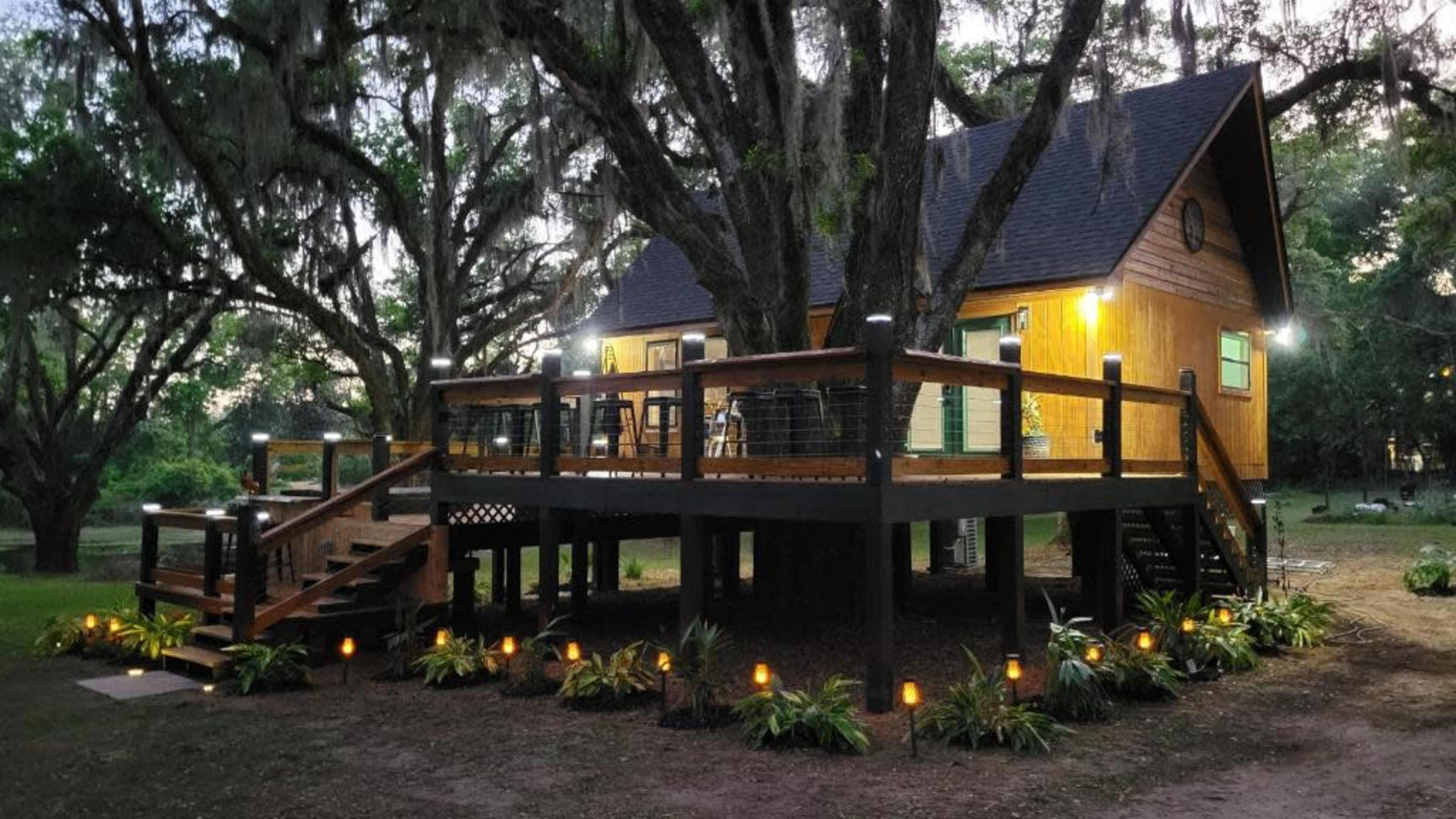 A wooden house on stilts with a large deck, surrounded by trees and garden lighting.