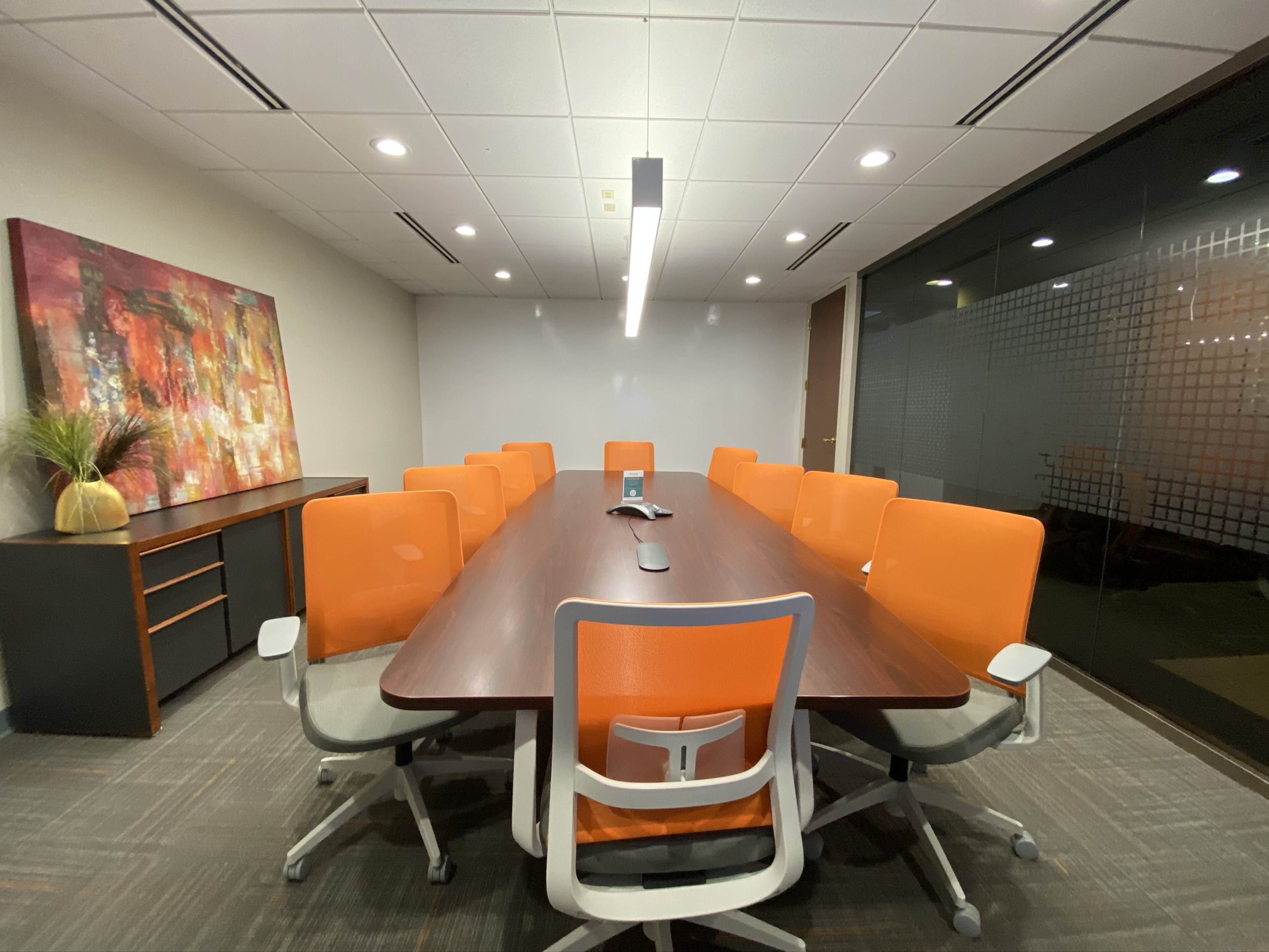 A conference room features a long wooden table surrounded by orange chairs, with a modern art piece and a plant on the wall.